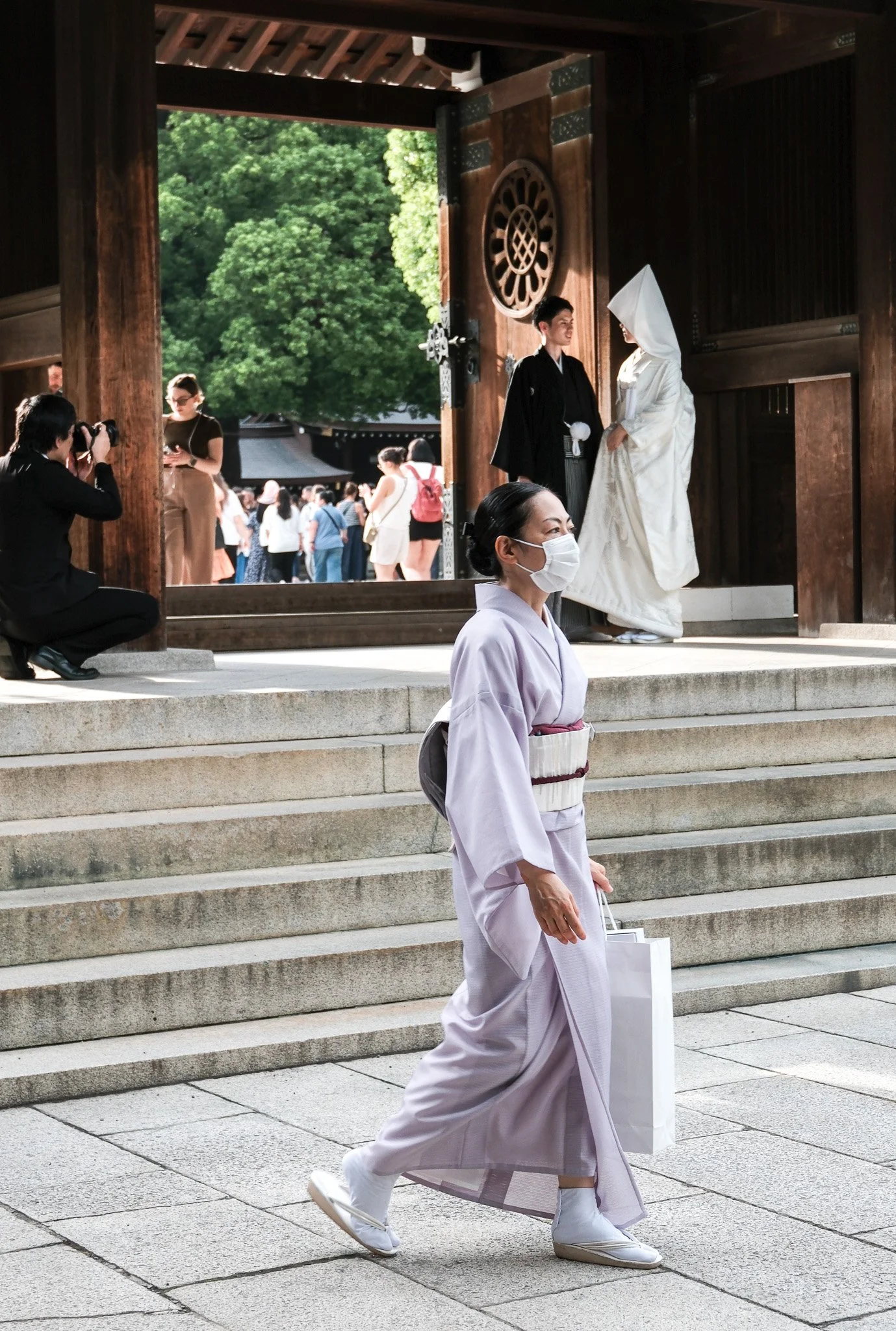 Eine Frau in traditioneller japanischer Kleidung, die eine weißen Kimono trägt, geht die Treppe hinunter, während eine Hochzeit im Hintergrund in einem Shinto-Schrein stattfindet.
