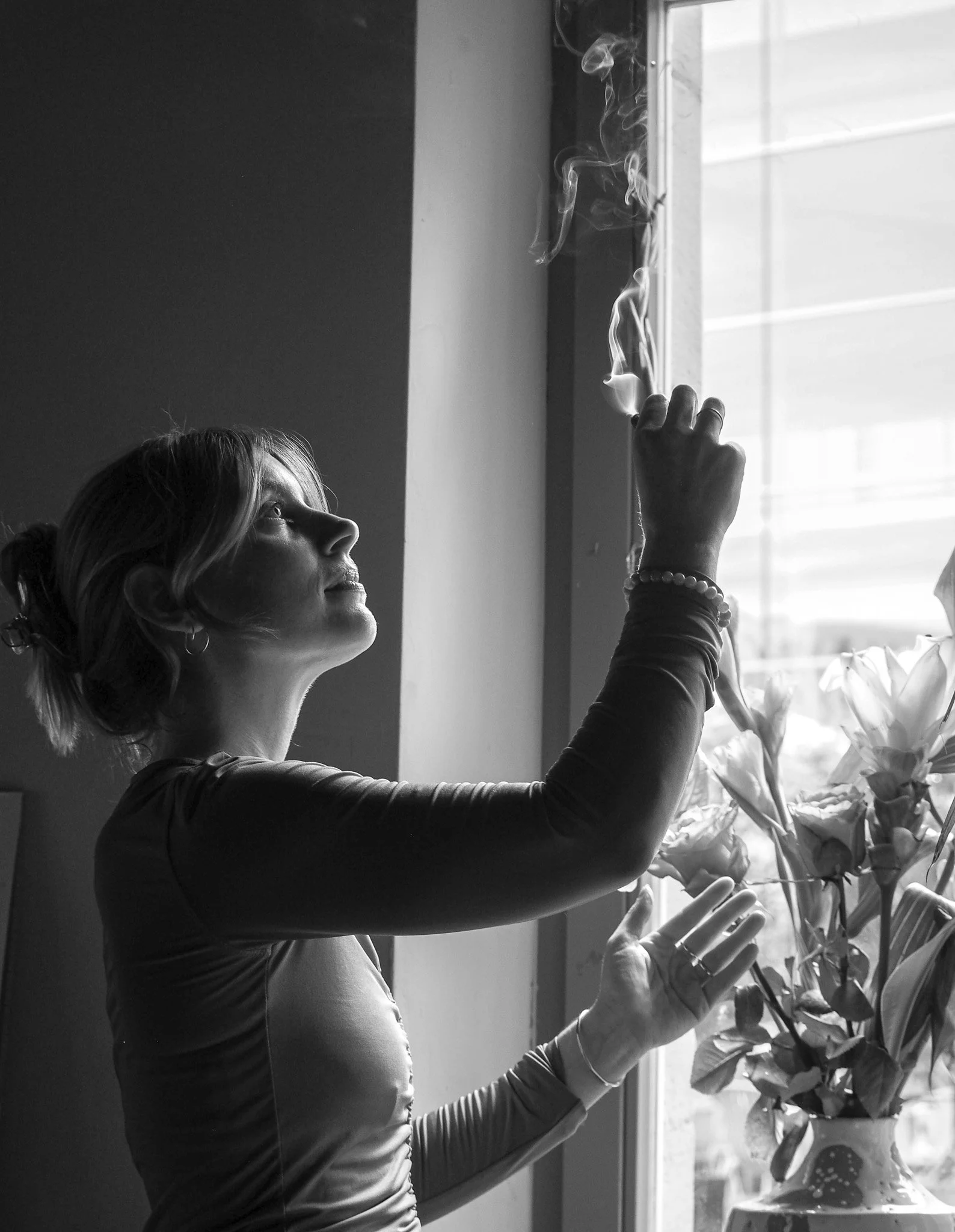 Schwarz-Weiß-Bild einer Frau, die in einem Raum neben einem Fenster Blumen in eine Vase stellen möchte.