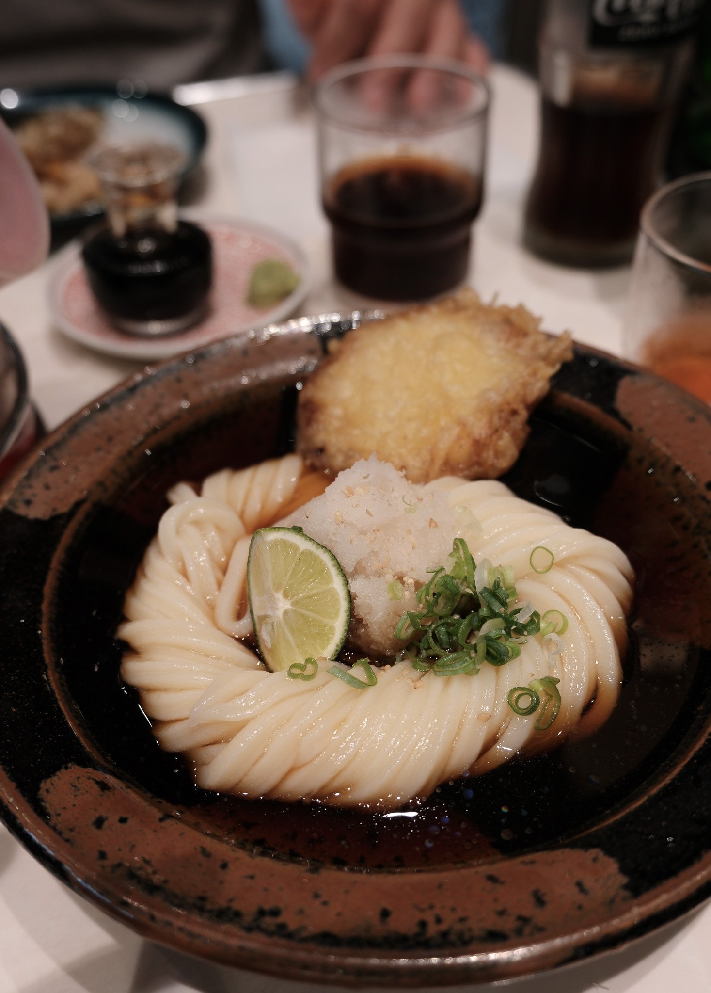 Eine Schüssel mit japanischer Udon-Nudel in Brühe, garniert mit Limettenscheibe, geriebener Daikon-Rettich, Frühlingszwiebeln und einem Stück Tempura.