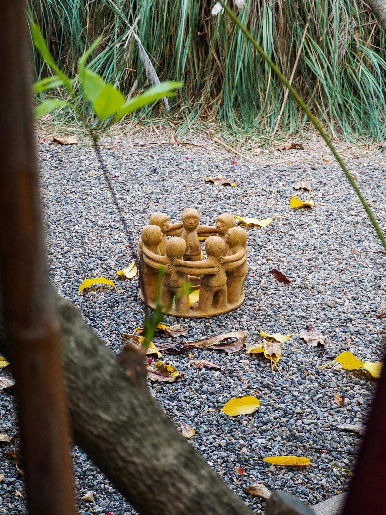Kunstvolle Figurengruppe aus Ton, die von Kindern in einem Kreis umarmt wird, auf einem Kiesweg mit Herbstlaub, umgeben von Pflanzen.