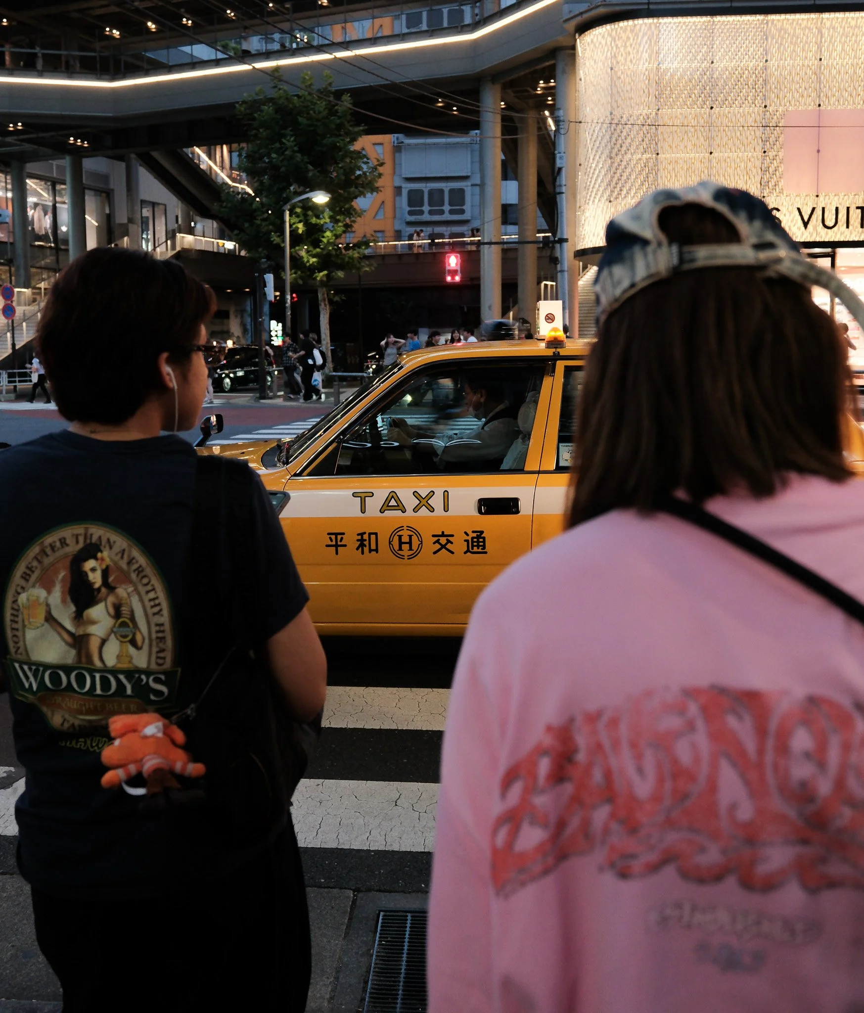 Zwei Frauen stehen an einer Straßenkreuzung in Japan, im Hintergrund ein gelber Taxi mit japanischer Beschriftung und Menschen, die die Straße überqueren, umgeben von modernen Gebäuden bei Dämmerung.