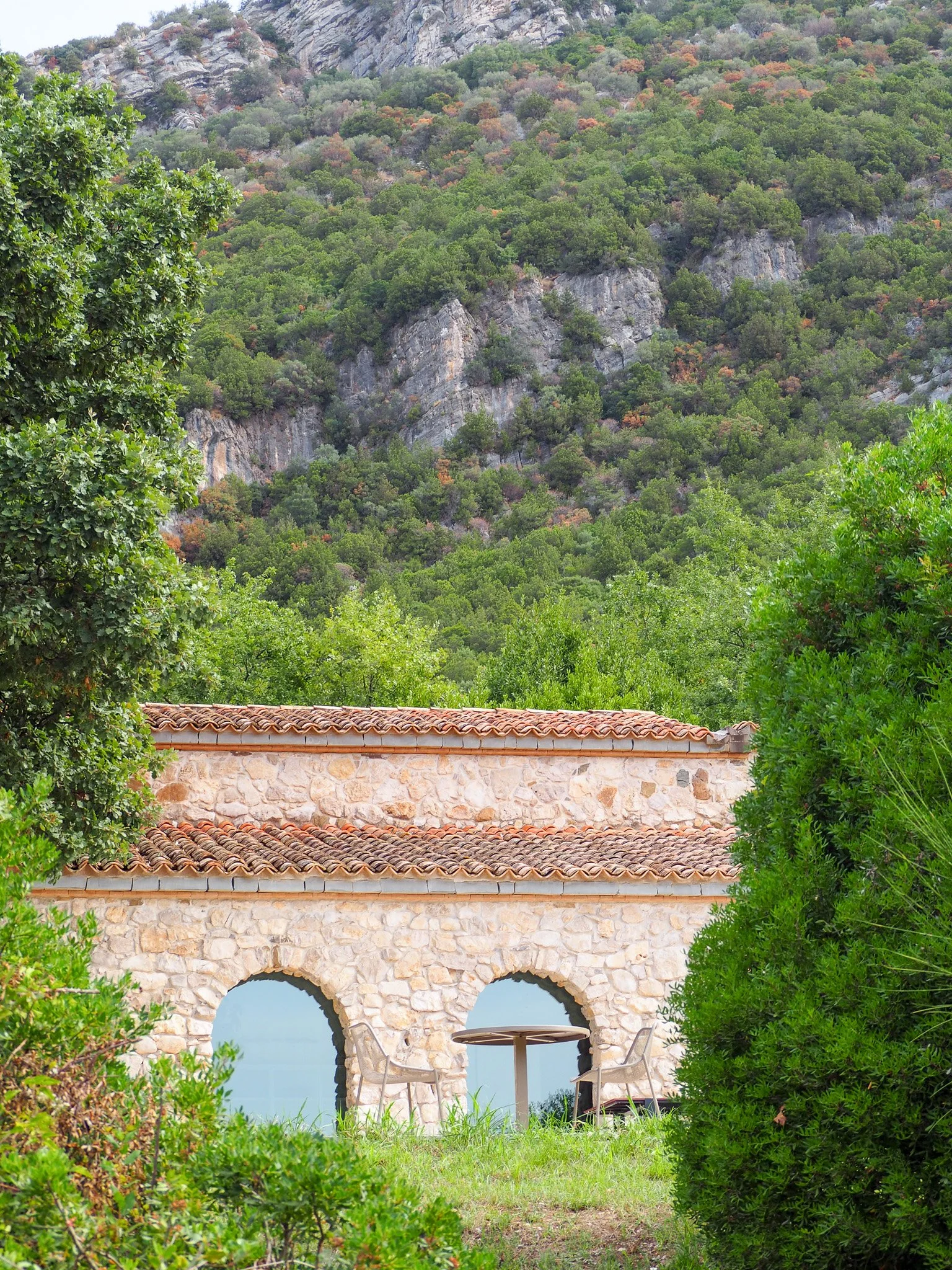 Ein Steinbau mit roten Ziegeldächern und zwei Bögen, umgeben von grünen Büschen und Bäumen, mit einem Tisch und zwei Stühlen davor, vor einer Berglandschaft mit dicht bewaldeten Hügeln.
