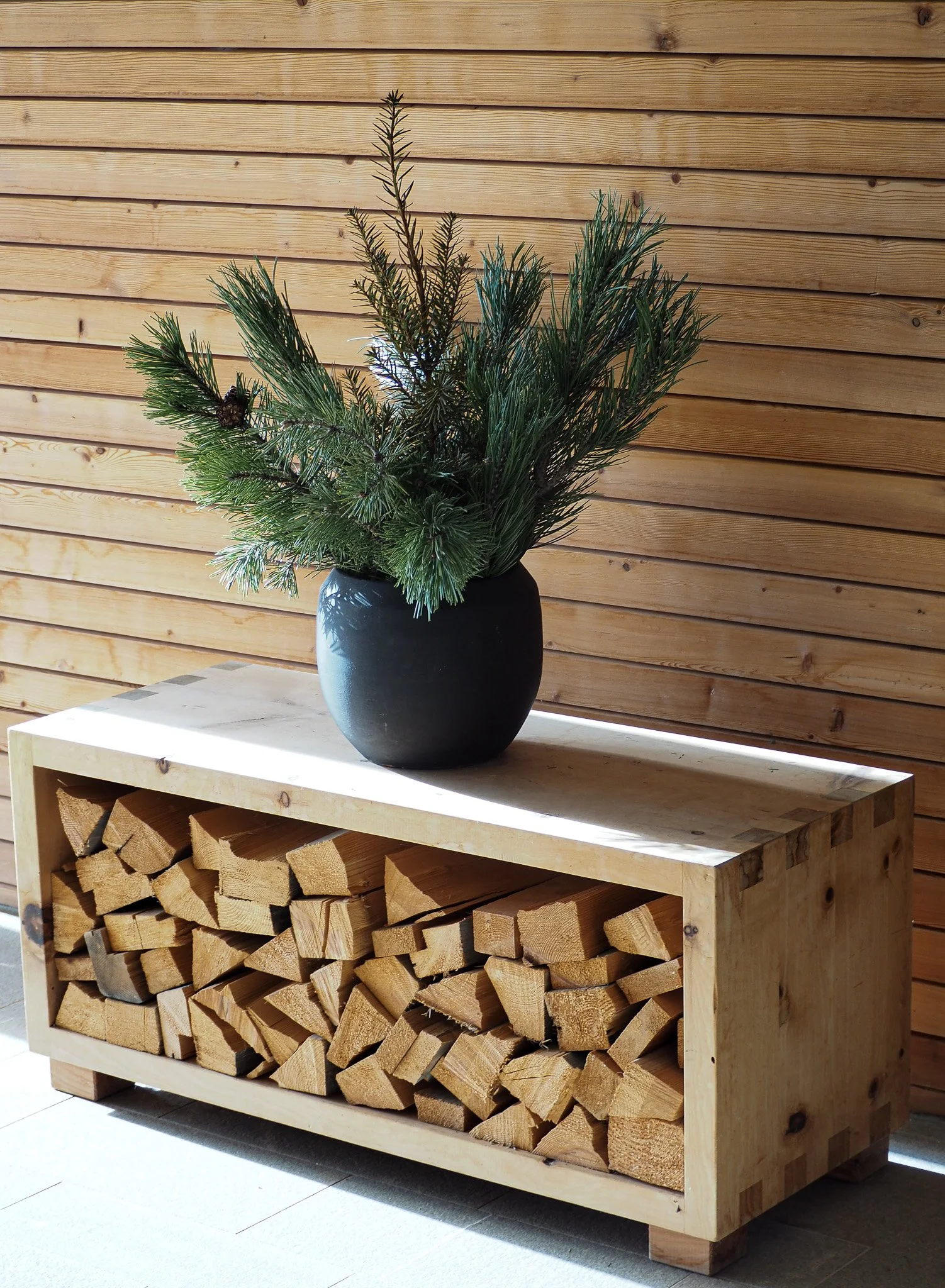 Ein Holzregal mit Brennholz, darüber eine schwarz-matte Vase mit Tannenzweigen auf einer Holzverkleidung.