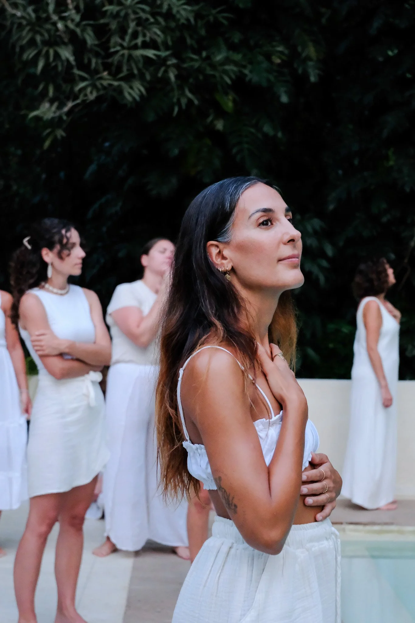 Eine Gruppe von Frauen in weißen Kleidern nimmt an einer zeremoniellen oder spirituellen Veranstaltung im Freien teil, die auf einer Terrasse vor einer grünen, bewaldeten Wand stattfindet.