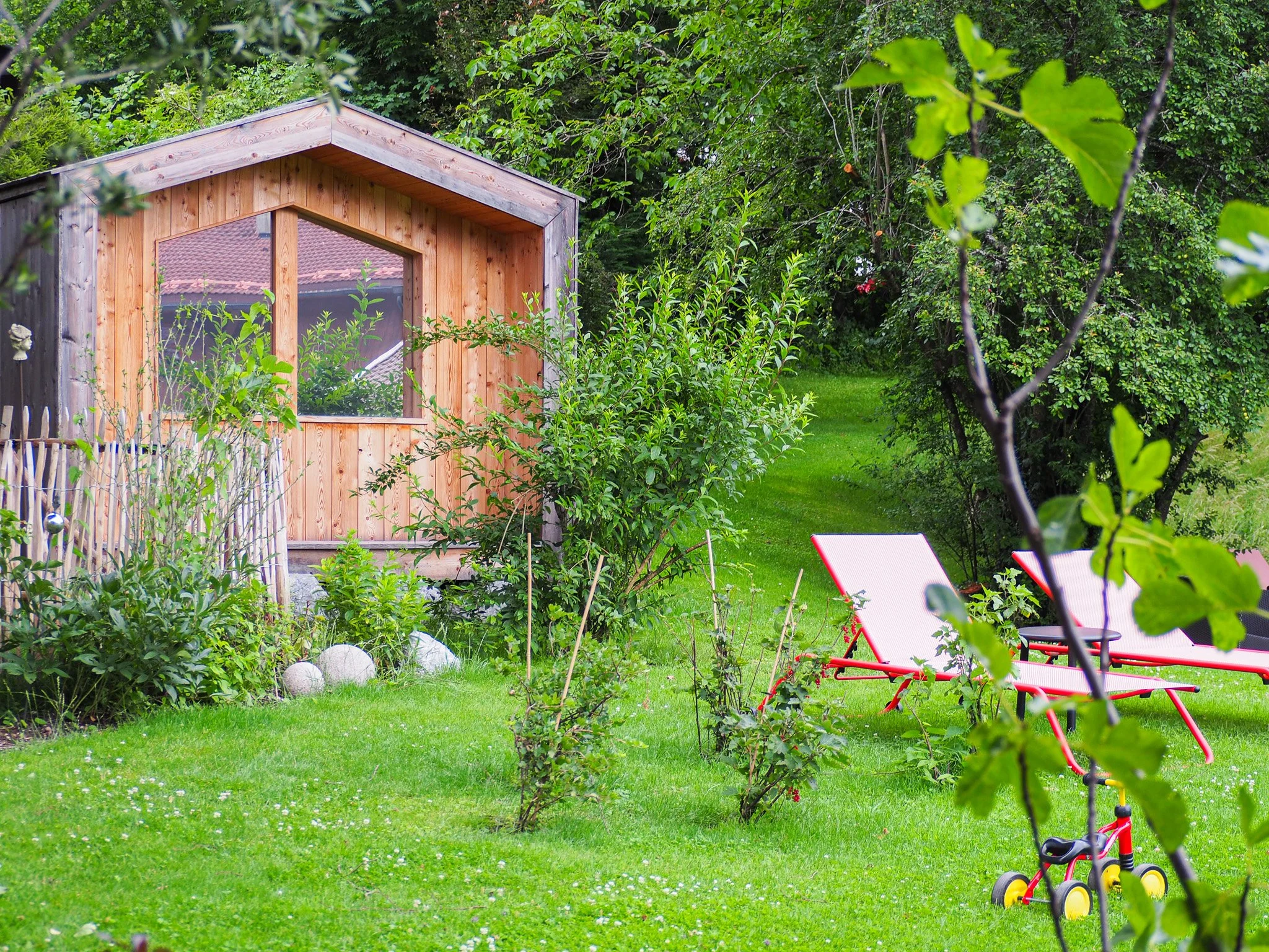 Garten mit kleiner Holzhütte, Grünflächen, Bänke, Kinderwagen und viele Bäume und Sträucher.