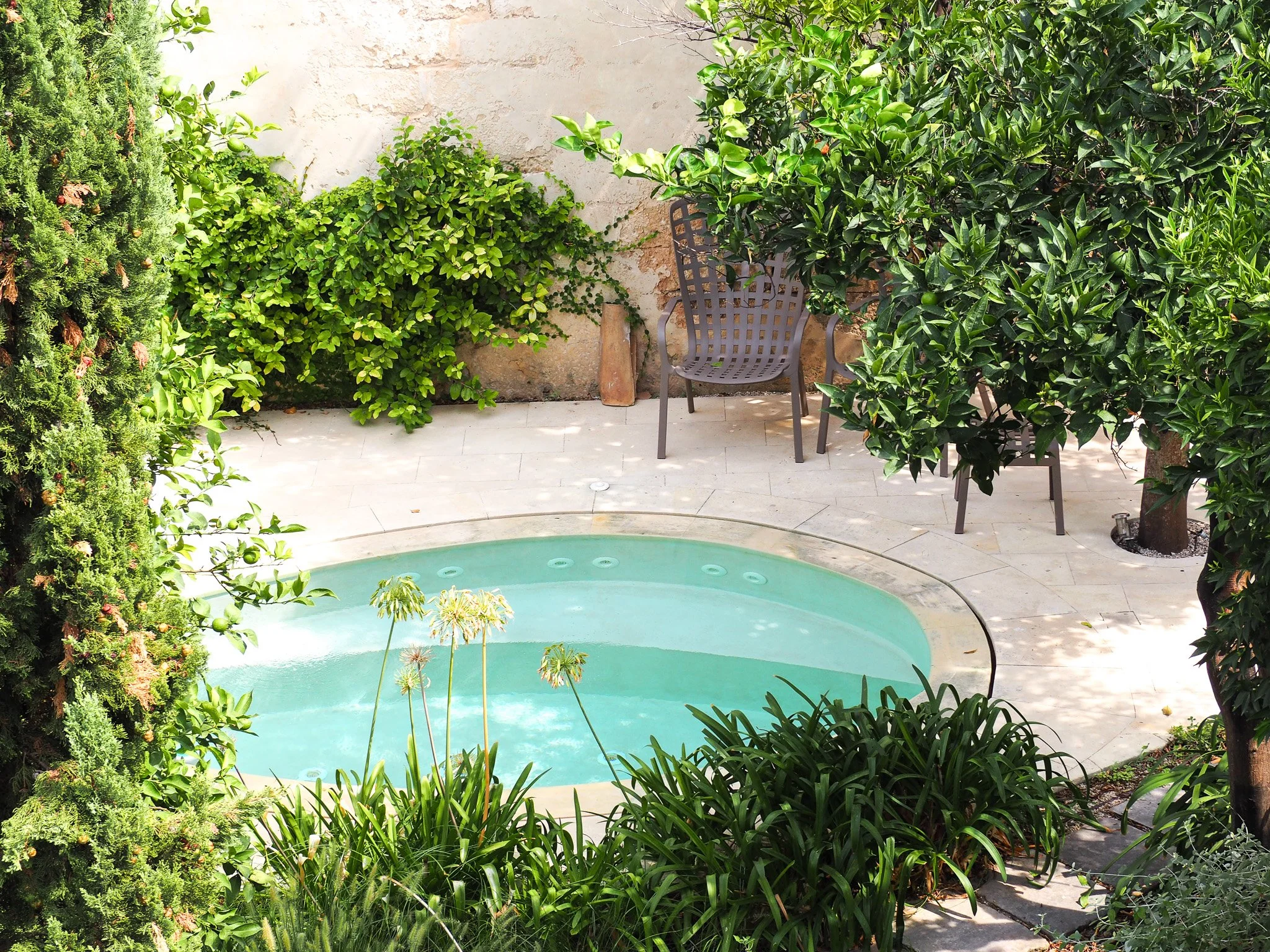 Ein kleiner Pool in einem Garten, umgeben von grünen Büschen und Bäumen, mit Gartenstühlen auf einem Steingarten.
