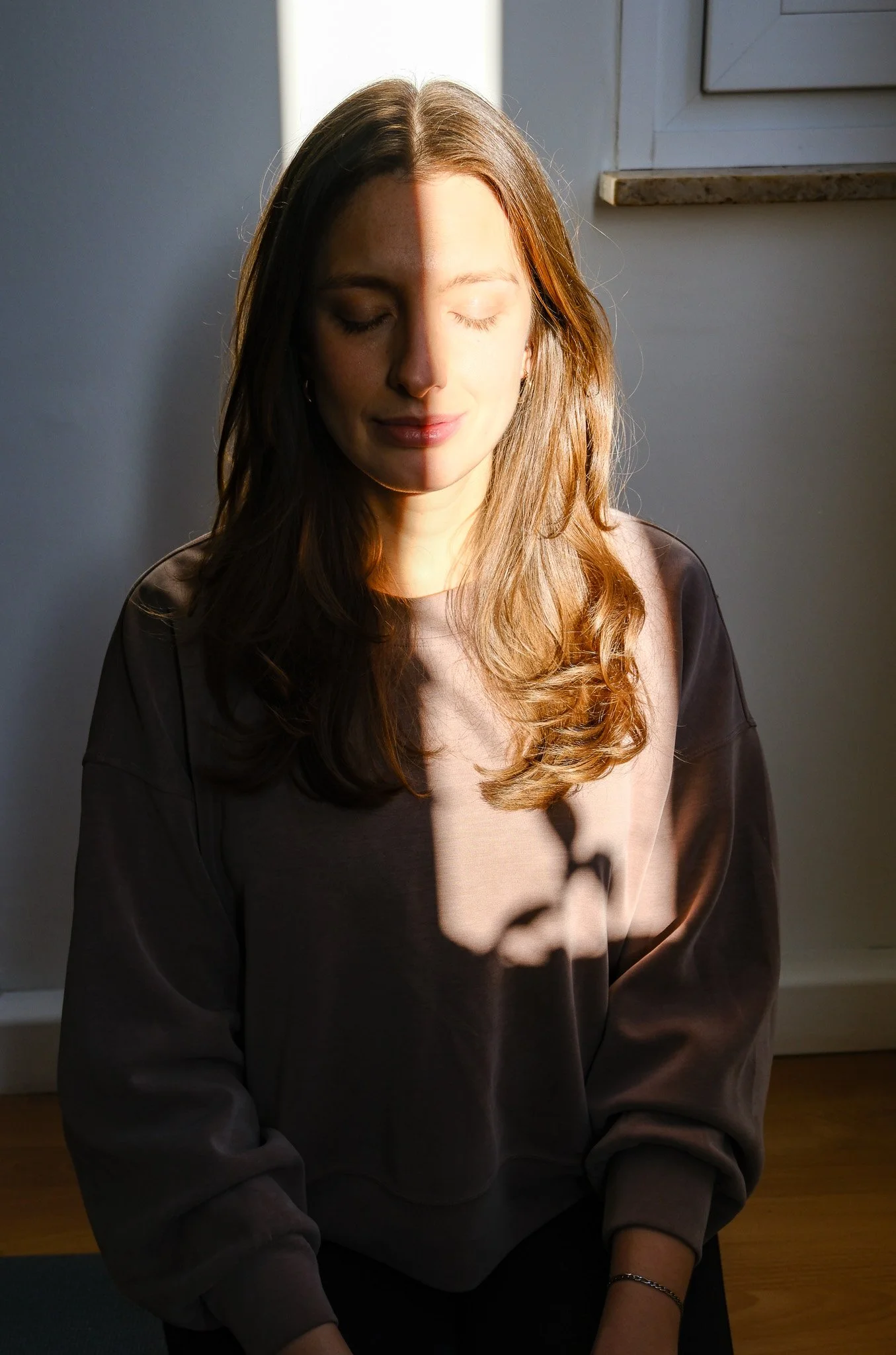 Junge Frau mit verschränkten Armen, die im Sonnenlicht sitzt, mit geschlossenen Augen und einem sanften Lächeln, im Hintergrund eine weiße Wand und Fenster