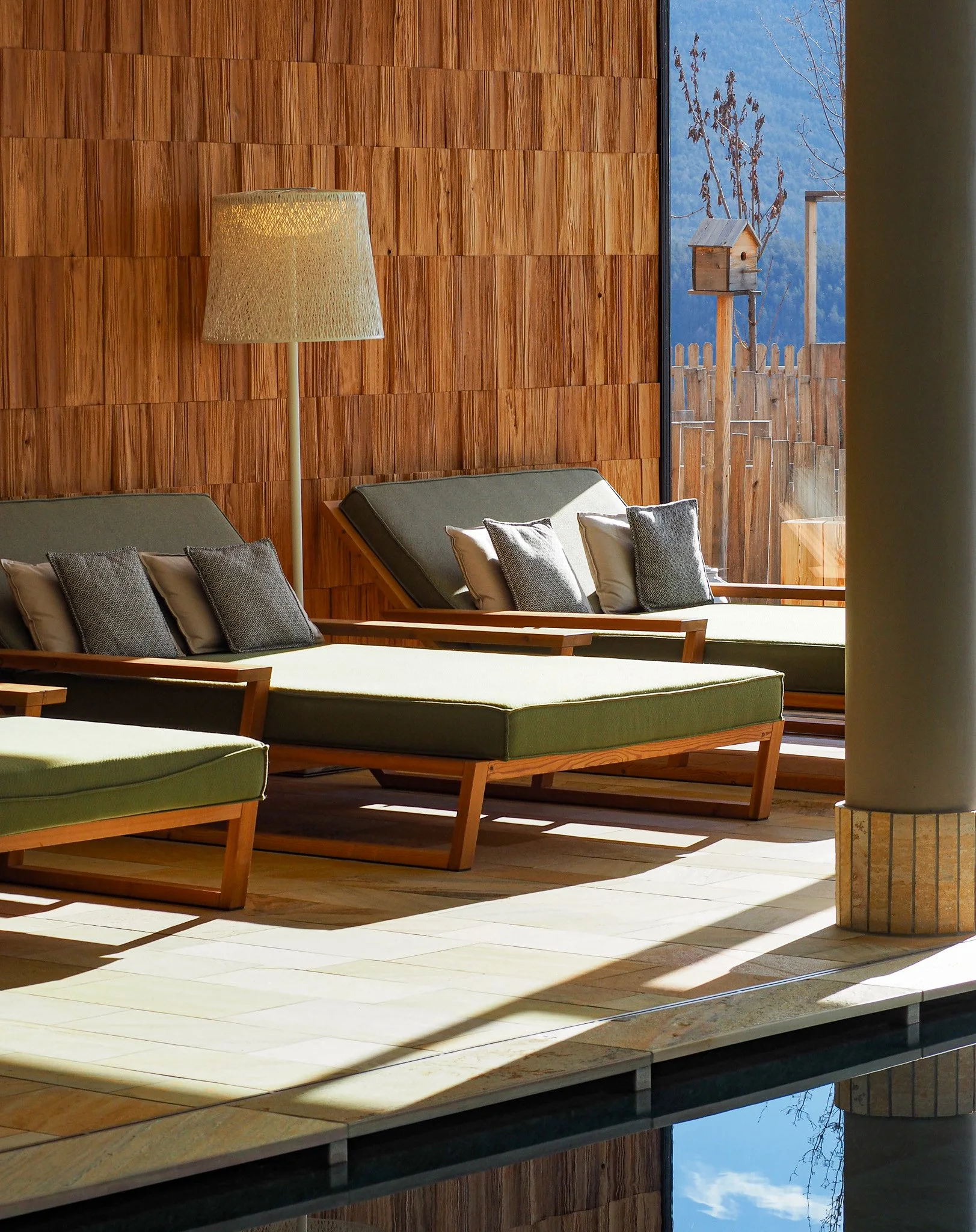 Sitzgelegenheiten mit grünen Polstern und grauen Kissen in einem Raum mit Holzverkleidung und Blick auf einen Garten mit Vogelhaus im Freien.