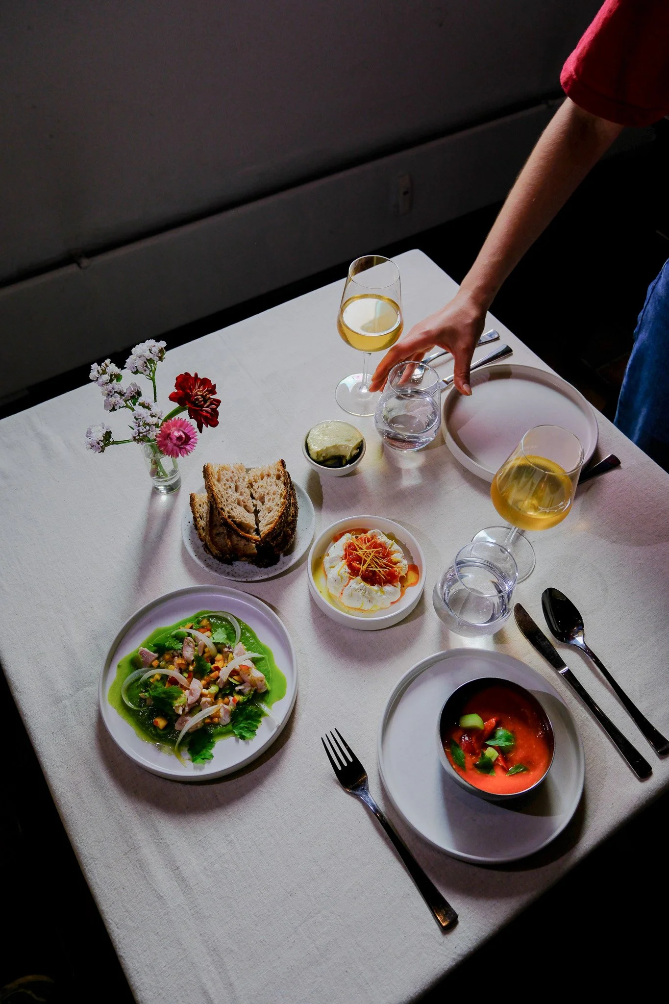 Ein gedeckter Tisch mit verschiedenen Speisen, zwei Gläser Wein, Wasser, Brot, eine kleine Vase mit Blumen und ein paar Besteck.
