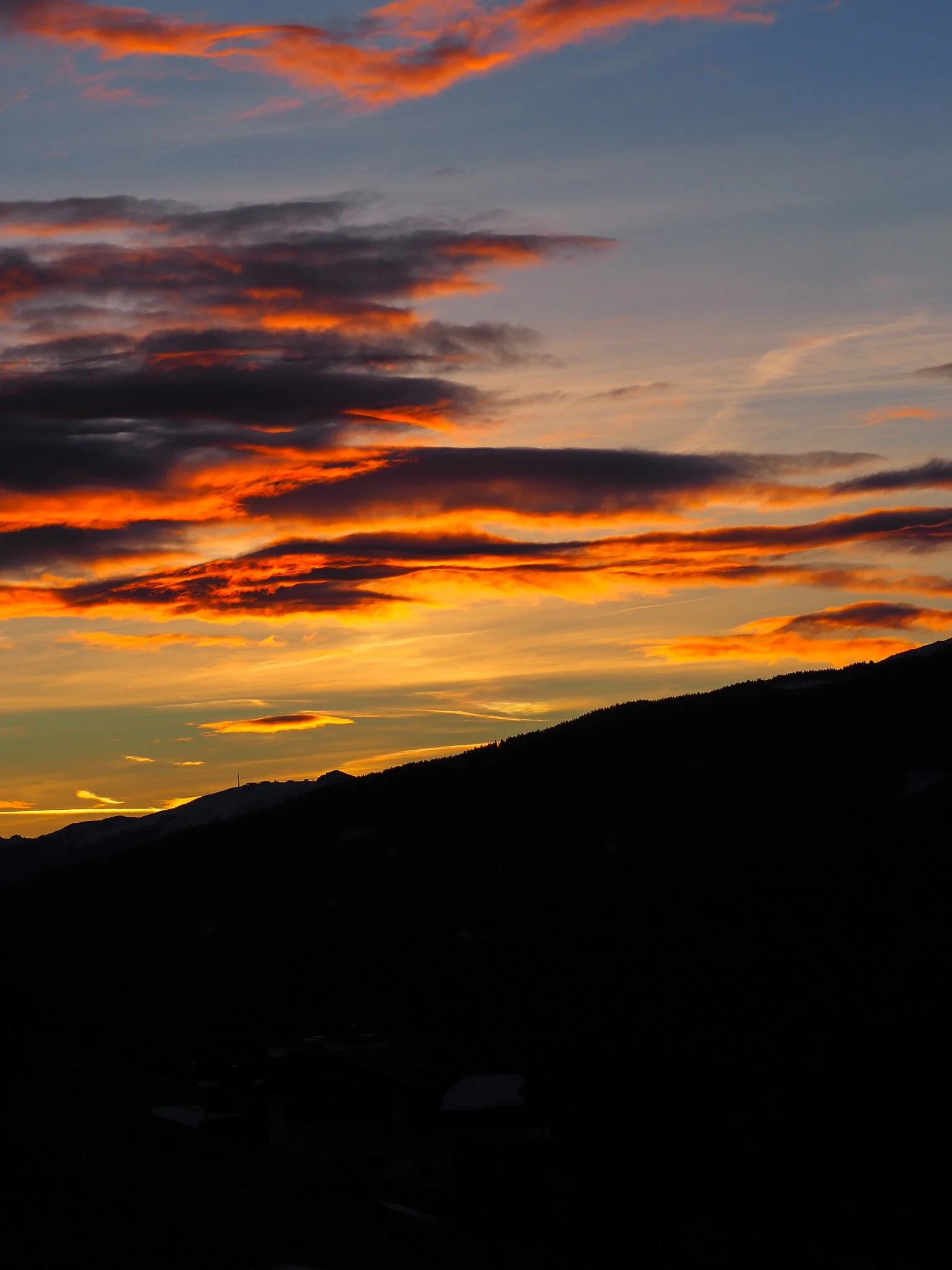 Ein farbenfroher Sonnenuntergang mit orange, gelben und blauen Wolken über einer dunklen Bergsilhouette.