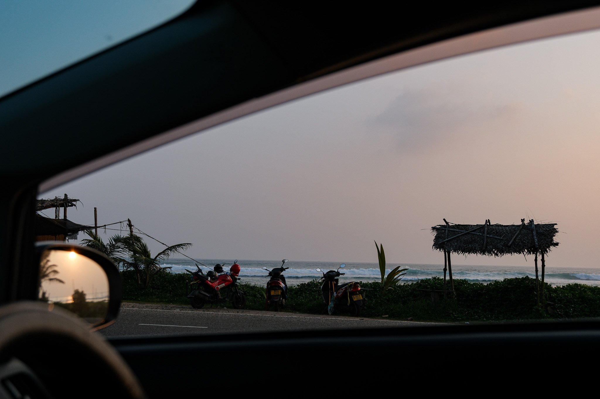 Blick aus einem Autofenster auf eine Strandlandschaft mit drei Mopeds, einem kleinen Strohhüttchen und Palmen bei Sonnenuntergang.