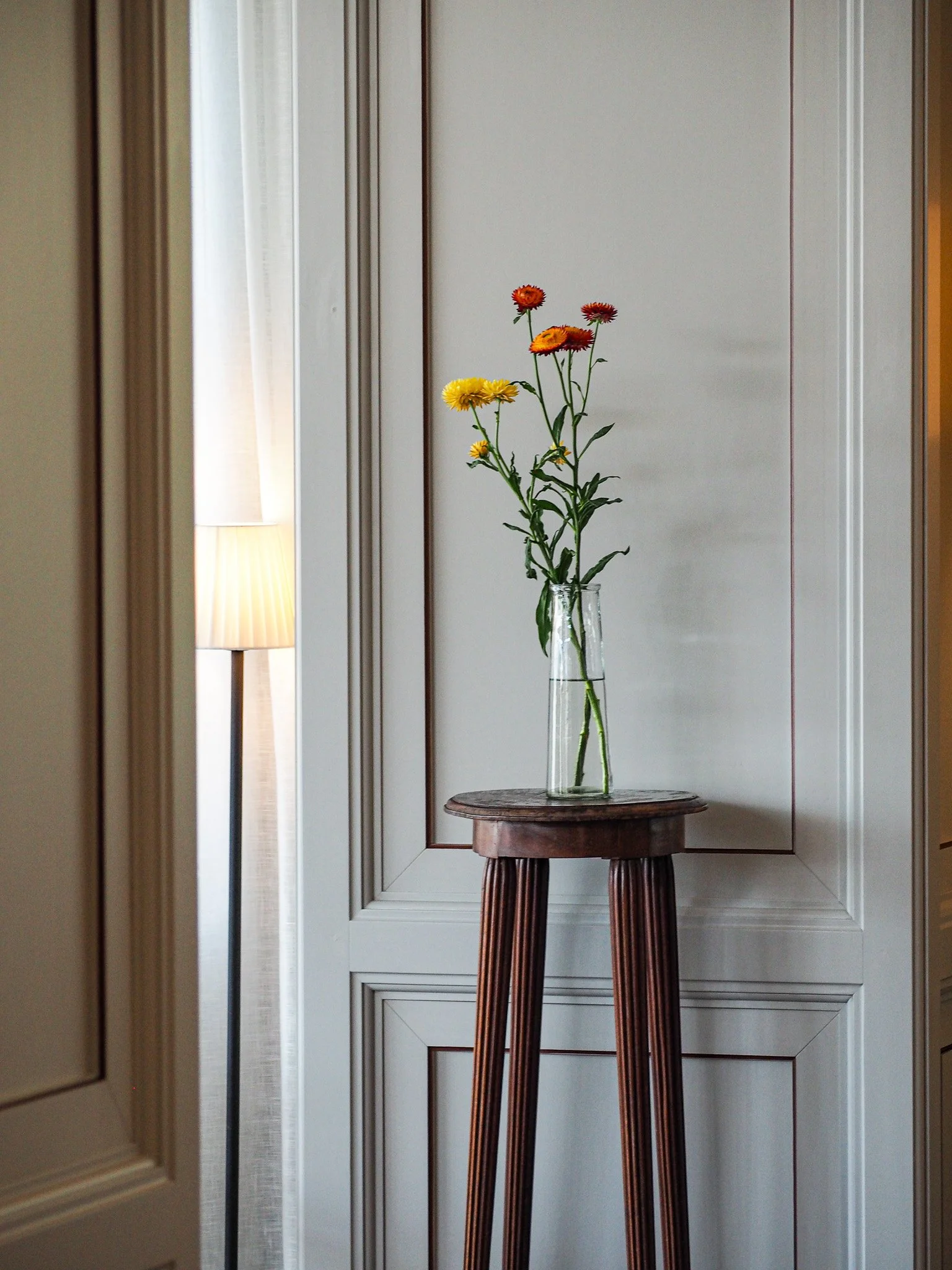 Bunte Blumen in einer Vase auf einem kleinen Holzhocker vor einer weißen Wand.