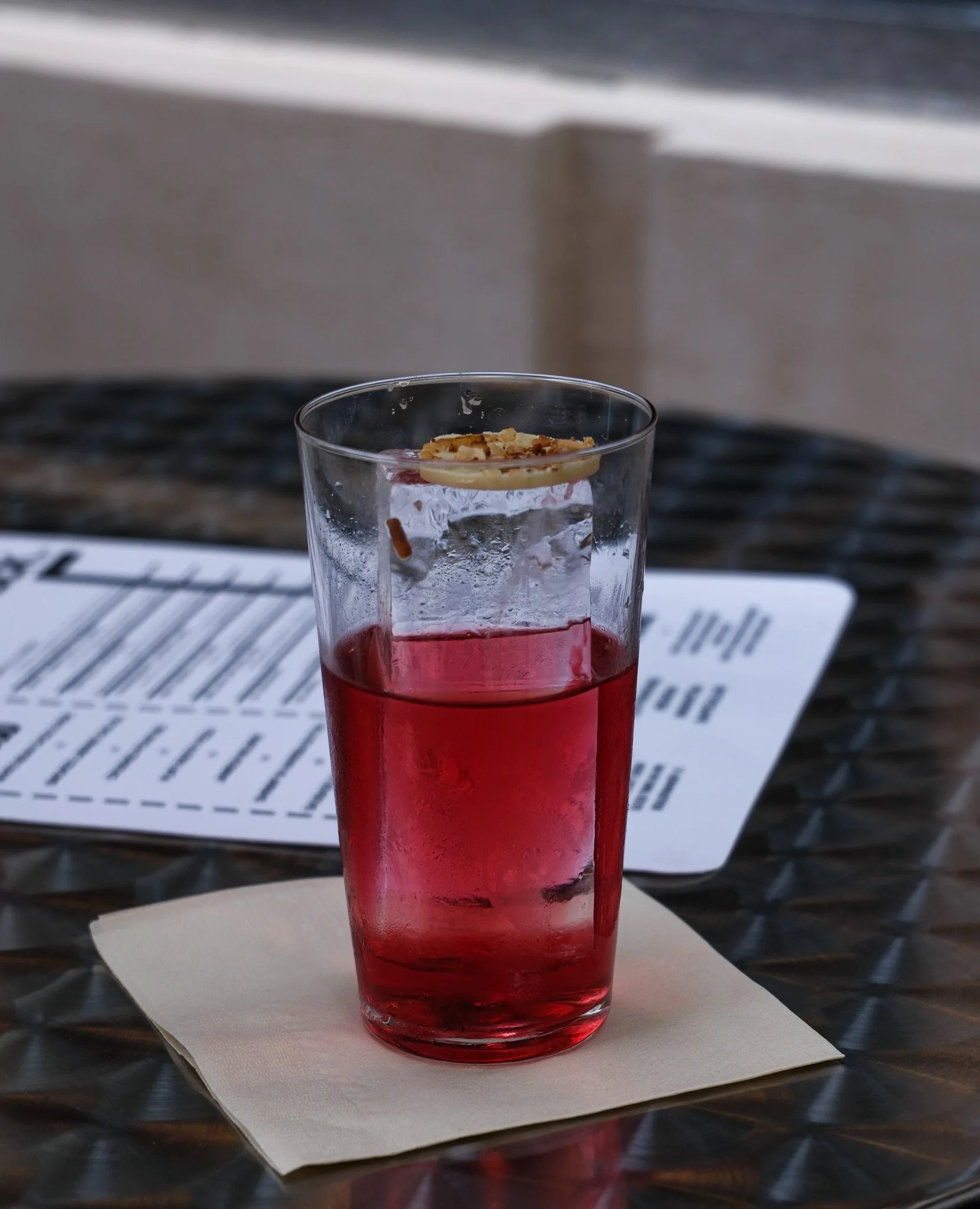 Ein hohes Glas mit rotem Getränk und einer Schicht aus Nüssen oder Schokolade oben drauf, auf einem Papieruntersetzer auf einem Tisch mit einem Menü im Hintergrund.