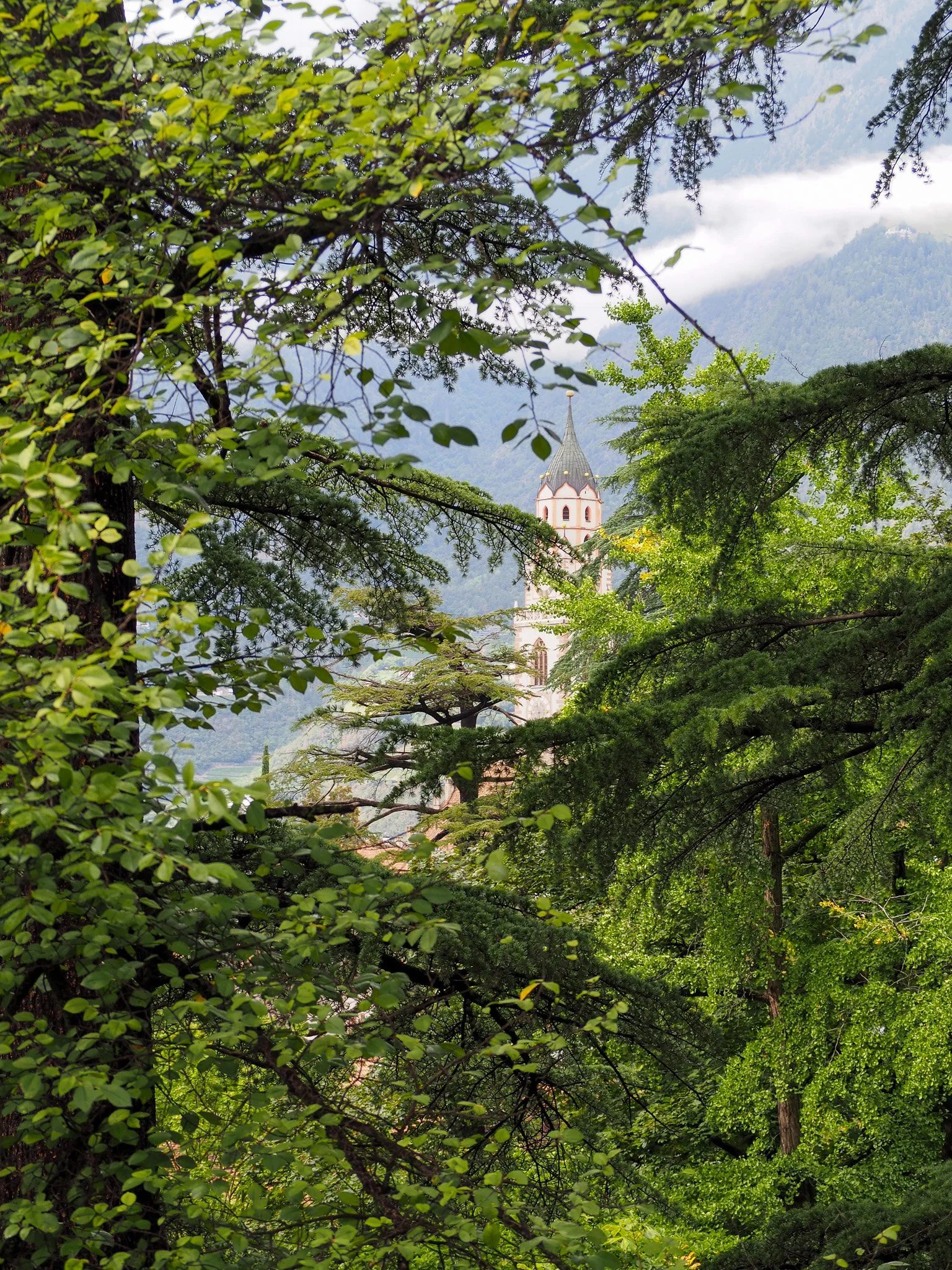 Blick auf einen Kirchturm, umgeben von grünen Bäumen in einer bergigen Landschaft.