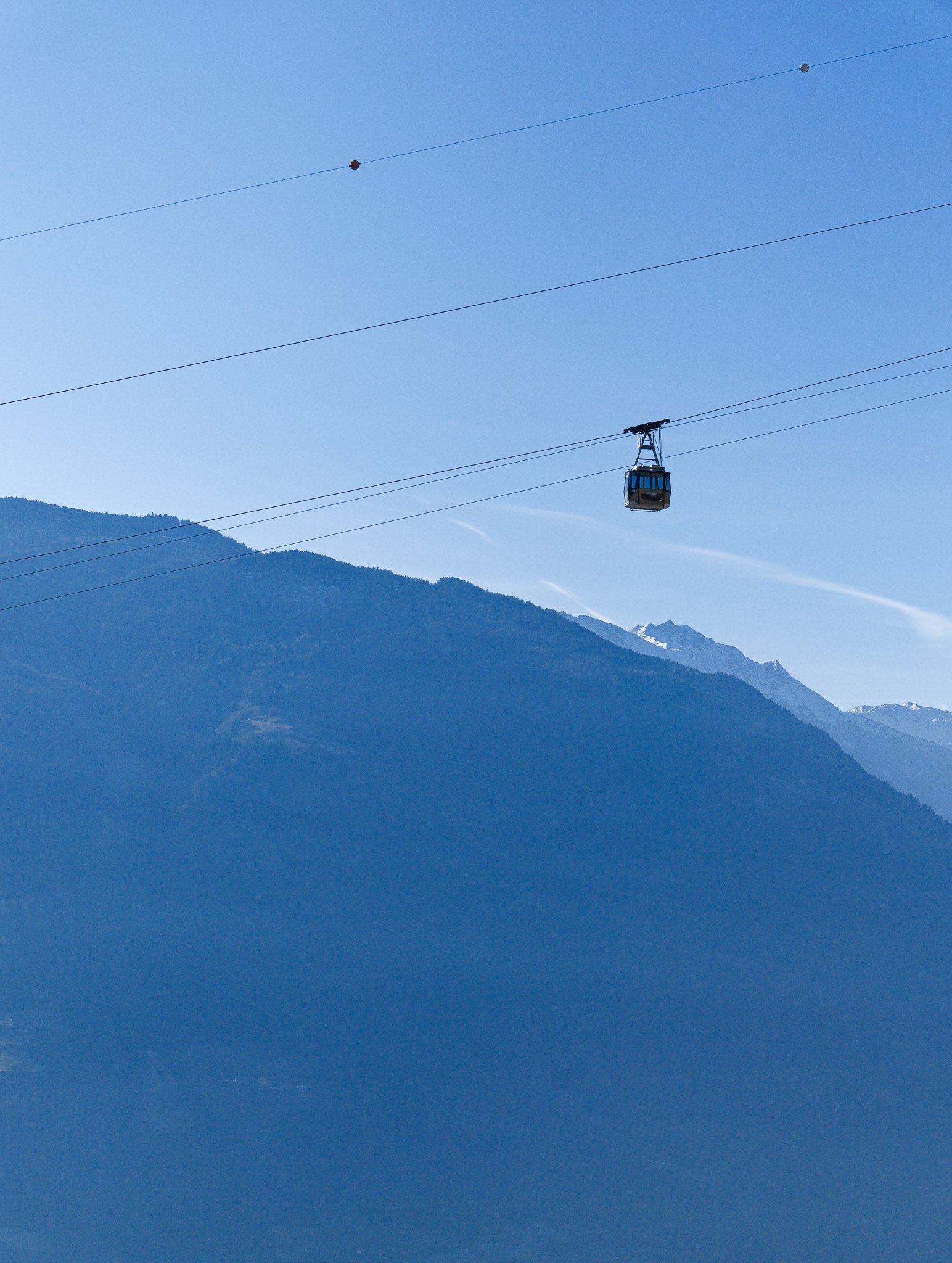 Eine Seilbahn fährt vor Berggipfeln mit Schneeresten in die Nähe. Der Himmel ist klar und blau.