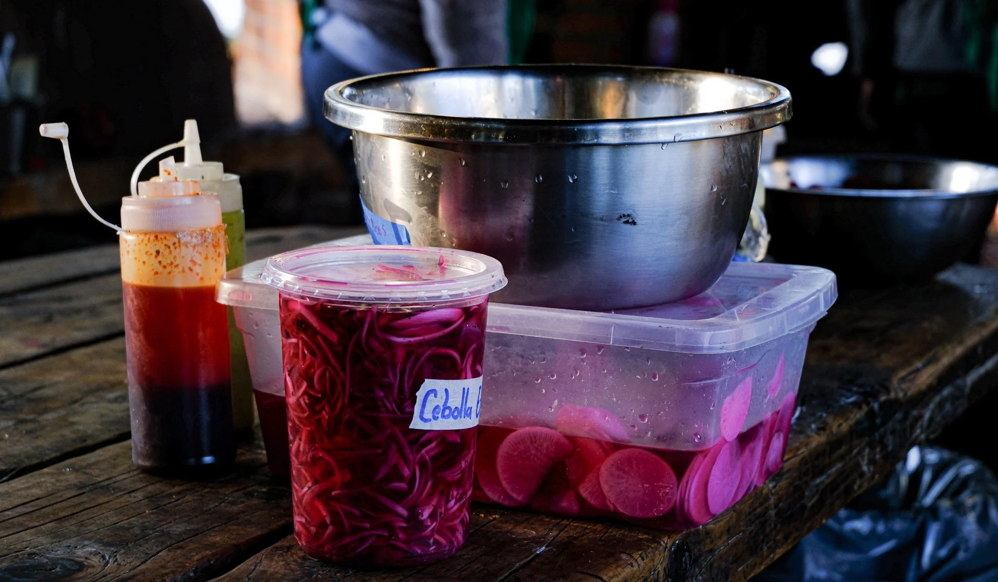 Lebensmittel und Küchenutensilien auf einem Holztisch, einschließlich einer Edelstahl-Schüssel, einem durchsichtigen Behälter mit roter Zwiebel, einer Flasche mit roter Sauce, einer Flasche mit grünlicher Flüssigkeit und einem schwarzen Behälter im H
