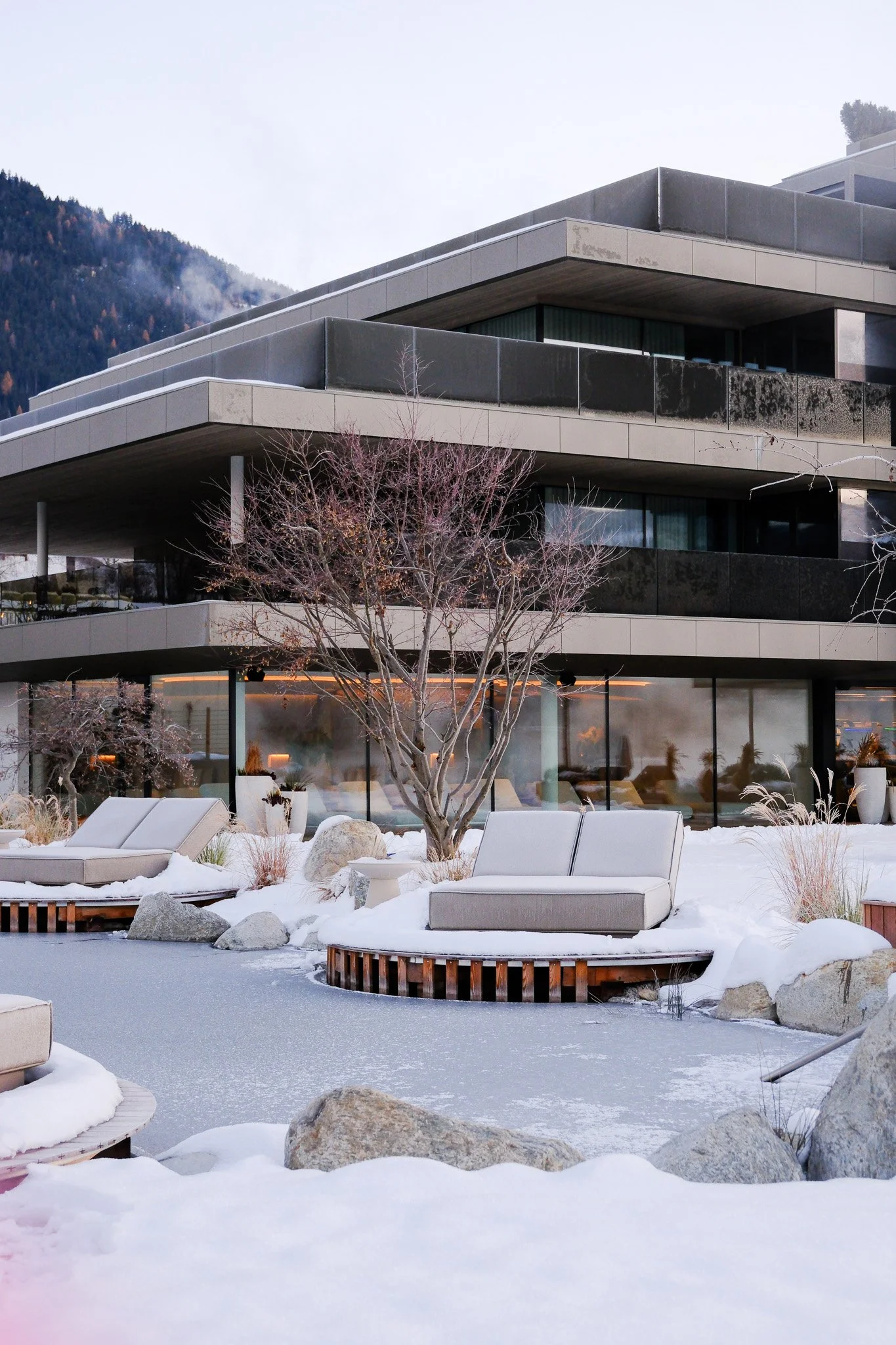 Modernes Gebäude mit großen Fenstern und Terrassen, umgeben von Schneelandschaft, gepflanzten Bäumen und Steinen, im Winter.