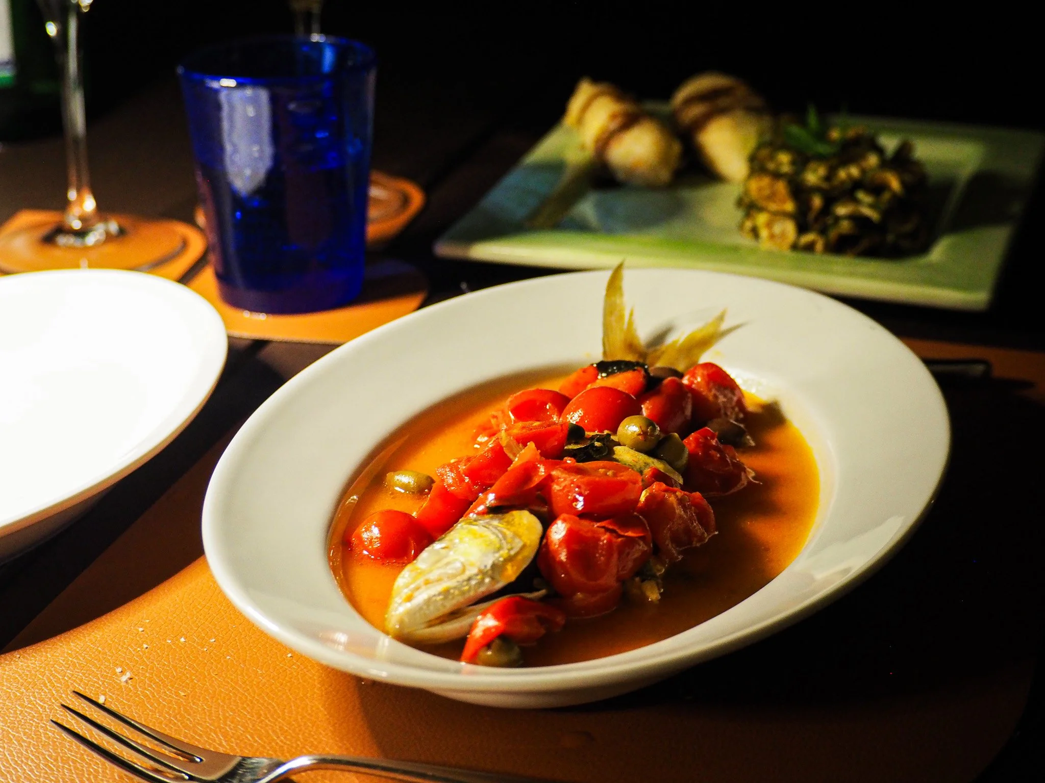 Fischgericht mit Tomaten, Paprika und Oliven in einer weißen Schüssel, Abendessen mit blauer Wasserglas, weiteren Tellern und Kerzen auf einem gedeckten Tisch.