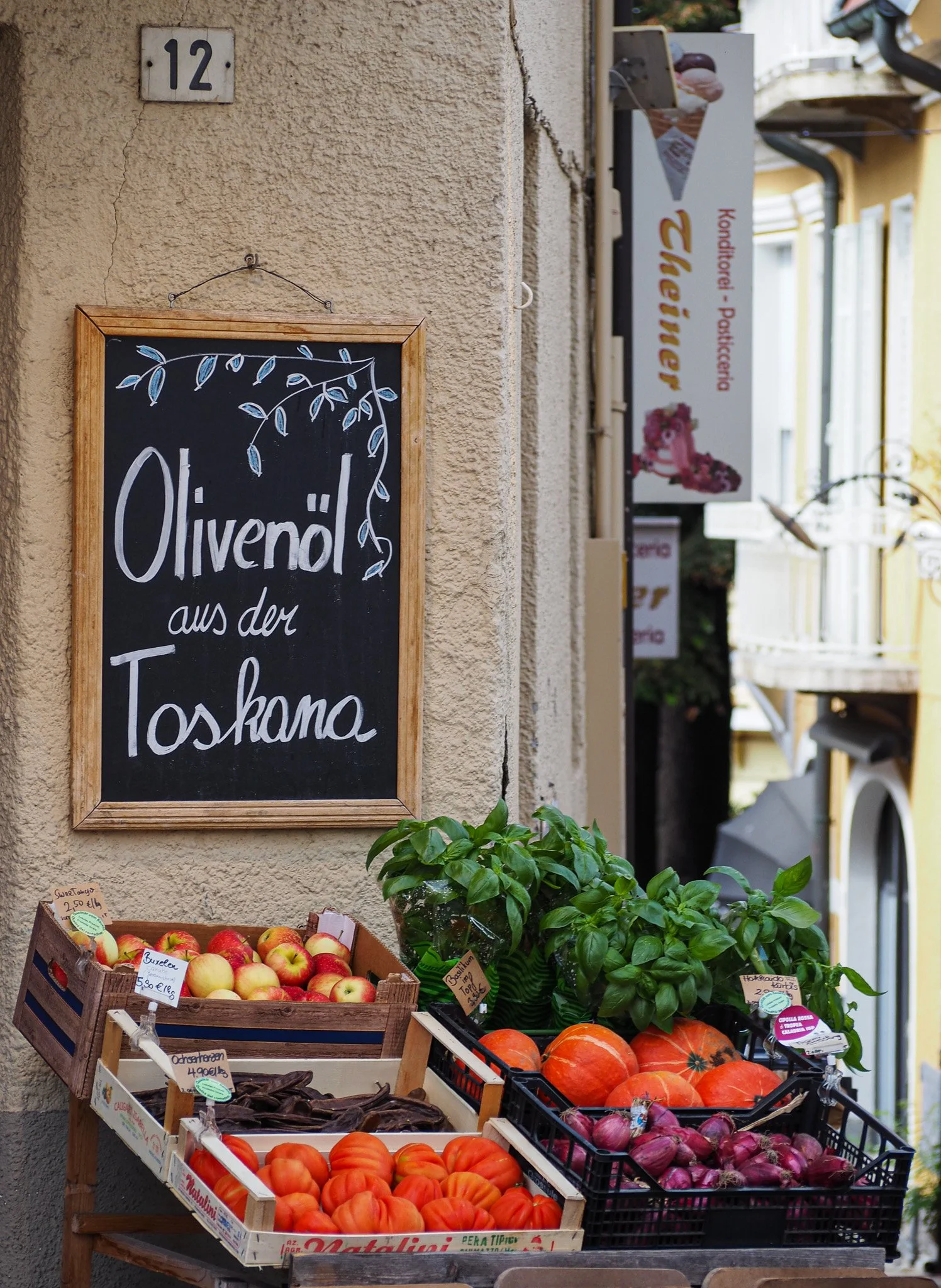 Obst- und Gemüsestände mit Äpfeln, Kürbissen, roten Zwiebeln, Basilikum und Tomaten vor einer Mauer auf einer Straße, Schiefertafel mit der Aufschrift 'Oliventrial aus der Toskana'.