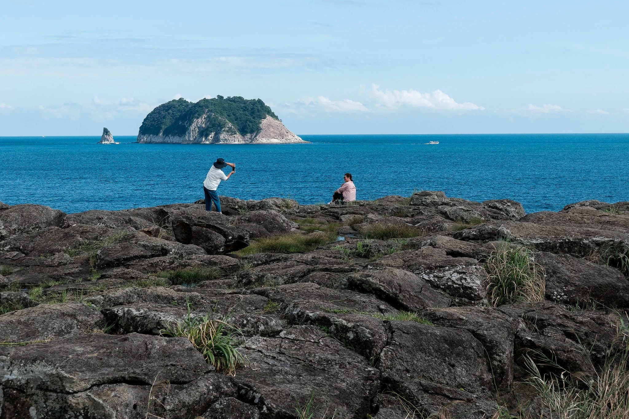Zwei Menschen auf einer Felsenpromenade am Meer, einer steht und macht ein Foto, der andere sitzt auf einem Felsen, im Hintergrund das Meer mit einer kleinen Insel und einem Boot.