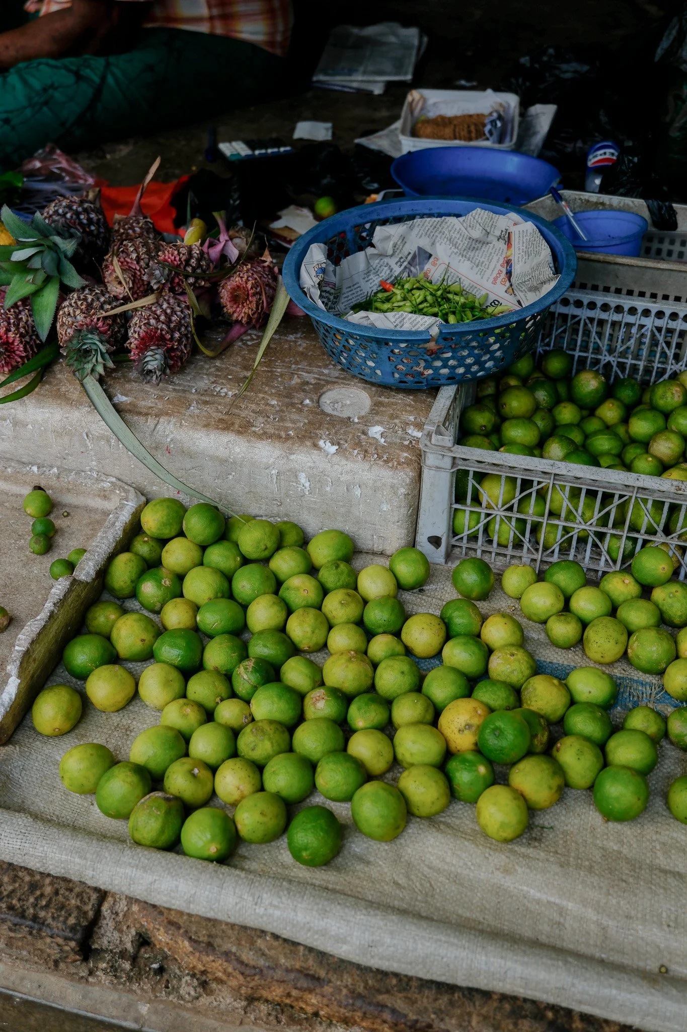 Verkaufsstand mit frischen Limetten, in einer Kiste und auf einem Tuch liegend, verbunden mit einem Marktstand, mit weiteren Limetten, Papier, und anderen Früchten und Gegenständen im Hintergrund.