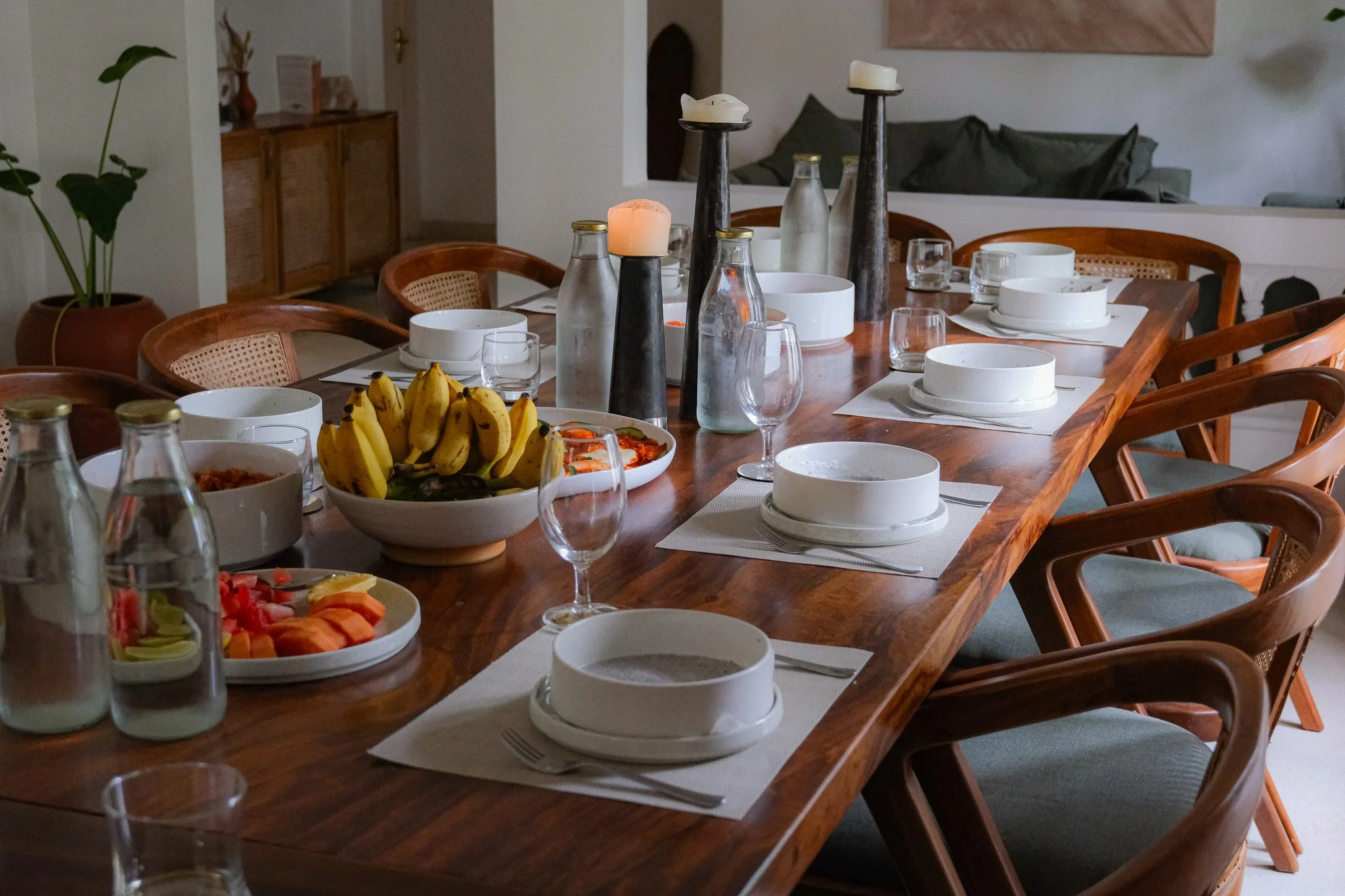 Ein gedeckter Esstisch mit weißen Tellern, Gläsern, Wasserflaschen, Kerzen, Früchten und einem Gericht. Im Hintergrund ein Wohnzimmer mit Sofa und Pflanzen.