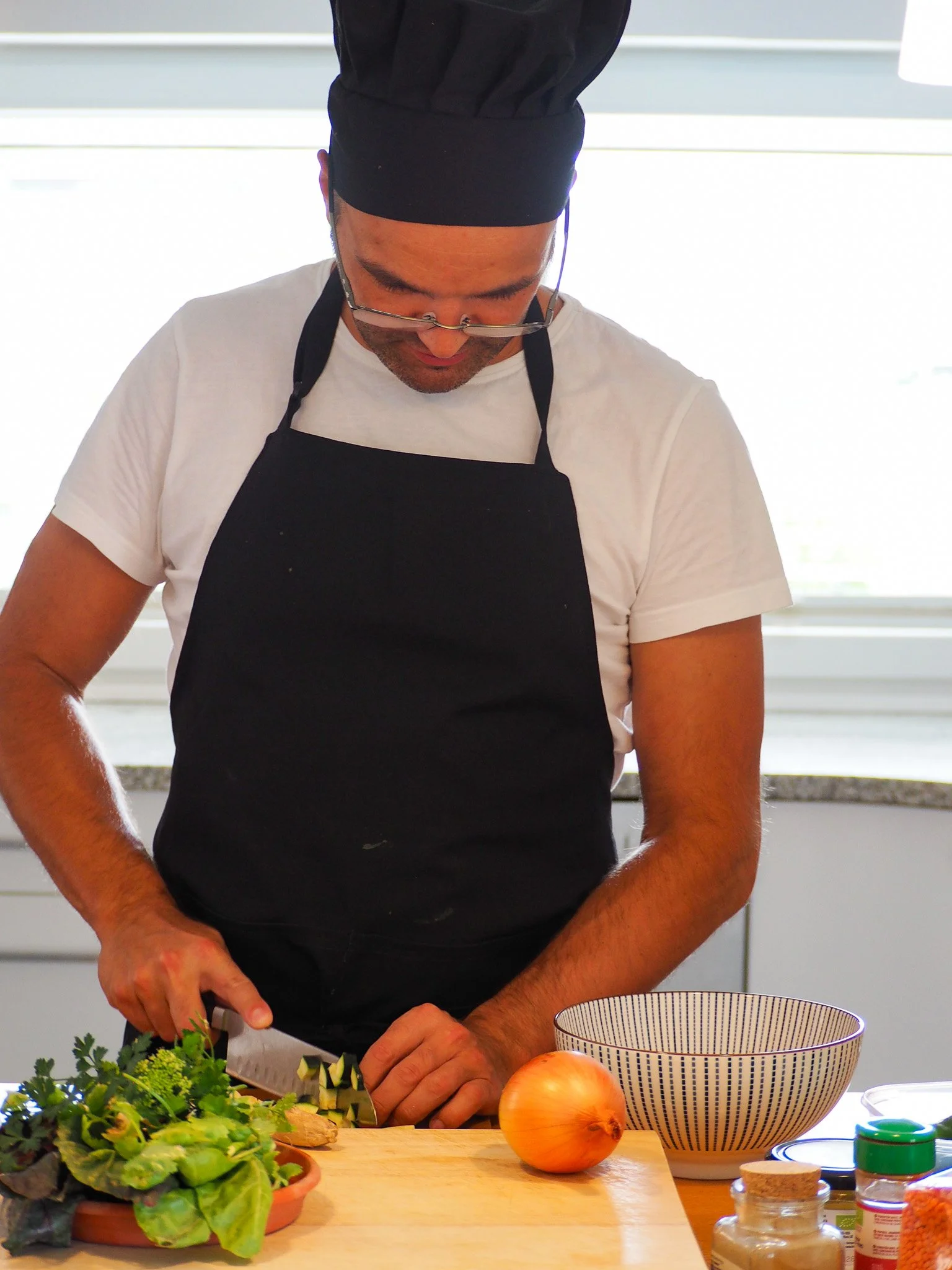 Ein Mann in einem schwarzen Kochhut und einer schwarzen Schürze schneidet Gemüse auf einer Küchenarbeitsplatte.