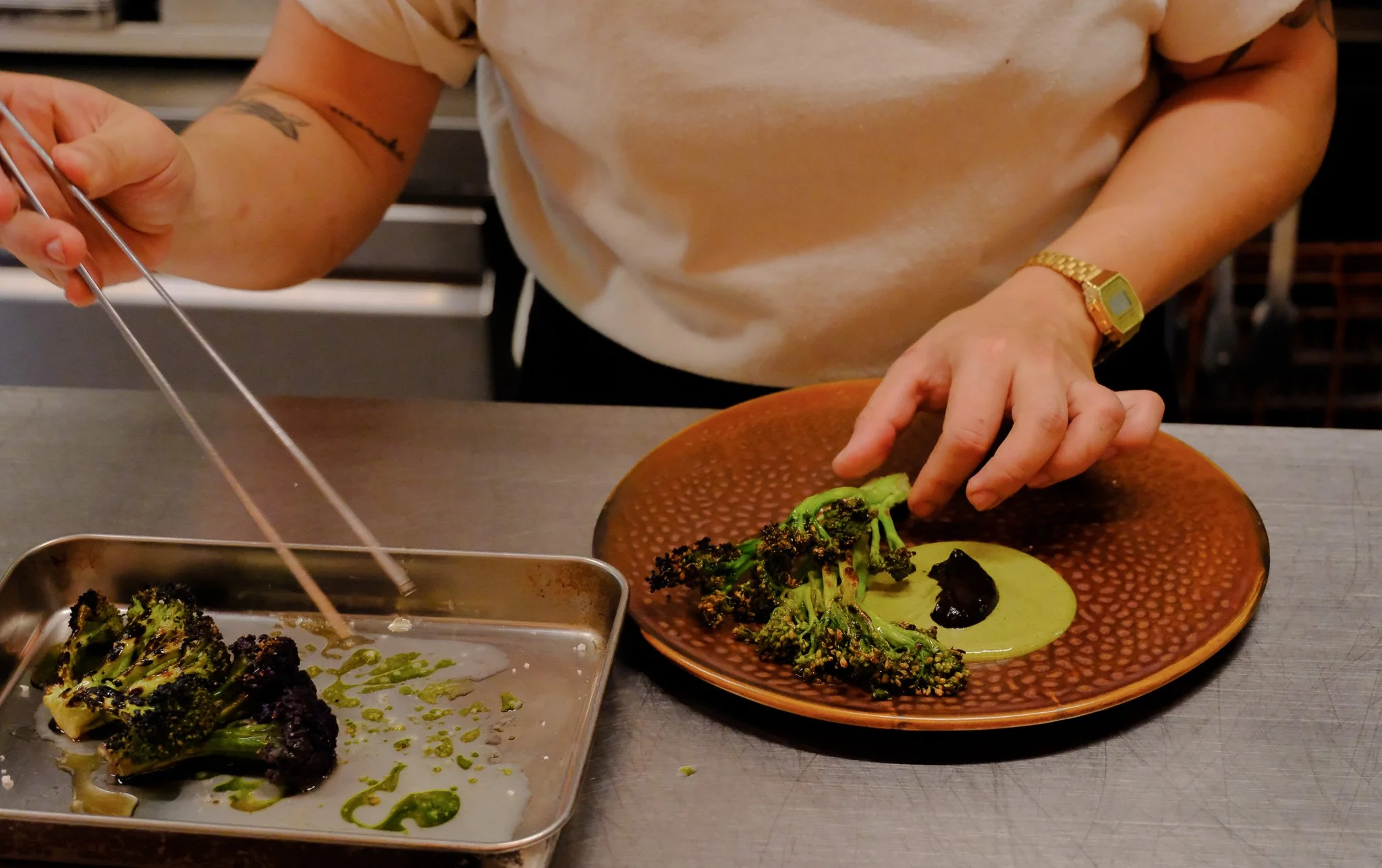 Person bereitet gegrilltes Brokkoli auf einem braunen Teller mit grünlicher Soße und schwarzer Deko. Die Person trägt eine goldene Armbanduhr und einen weißen T-Shirt, die auf einem grauen Arbeitsfläche arbeitet.
