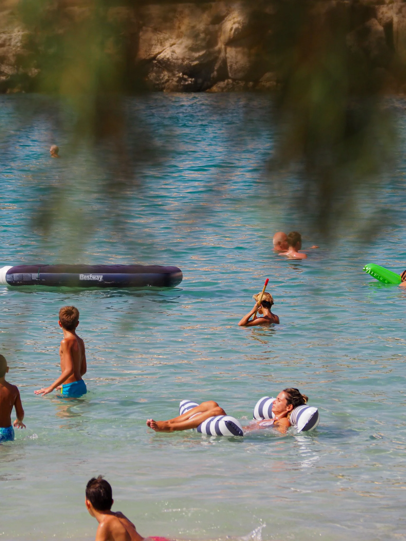 Menschen schwimmen und entspannen im Wasser an einem sonnigen Tag, einige mit Wasserspielzeug, im Hintergrund eine felsige Küste.
