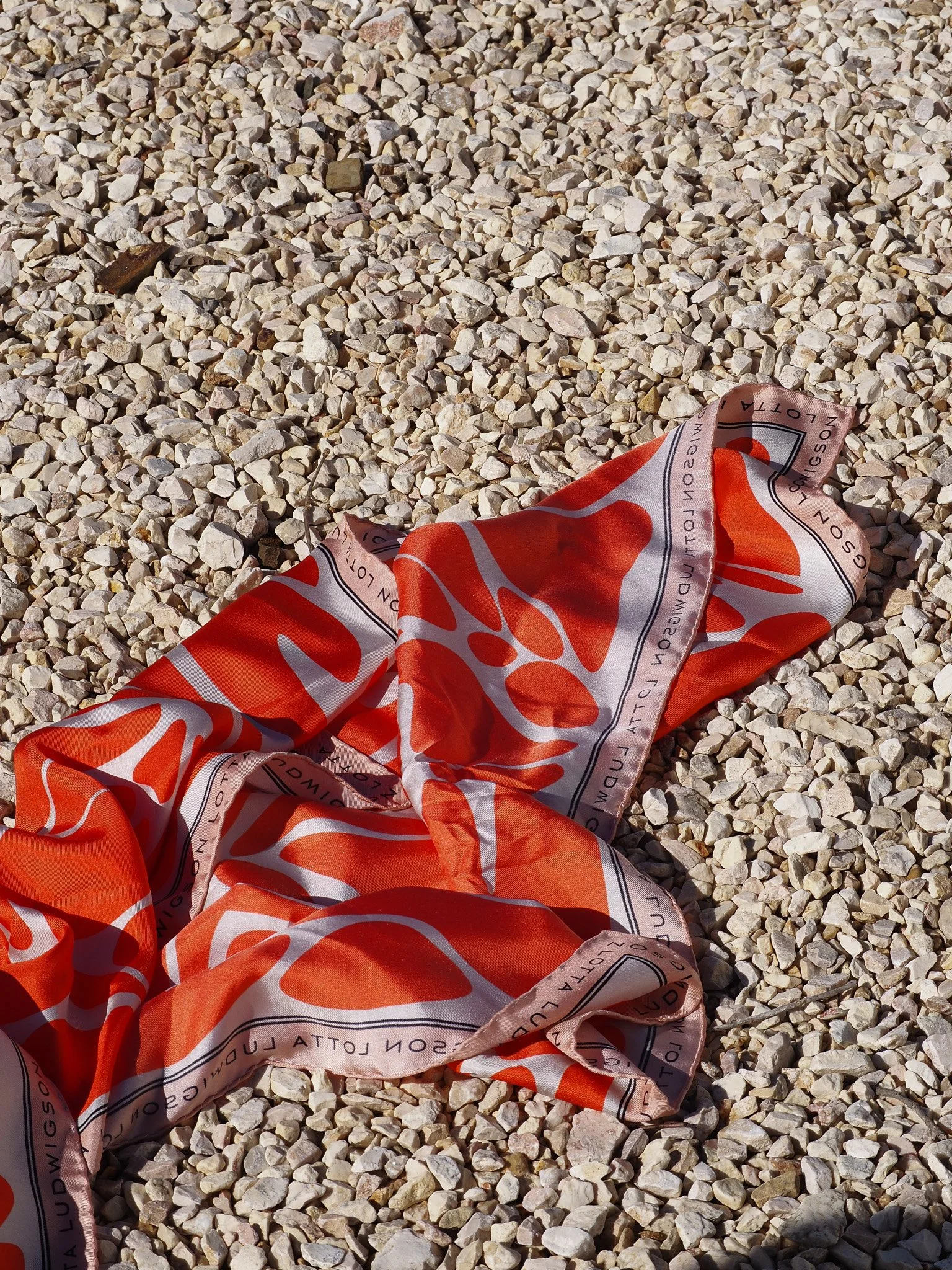 Ein zerknülltes rotes Seidentuch mit weißen und schwarzen Mustern liegt auf einem Kieselsteinboden.