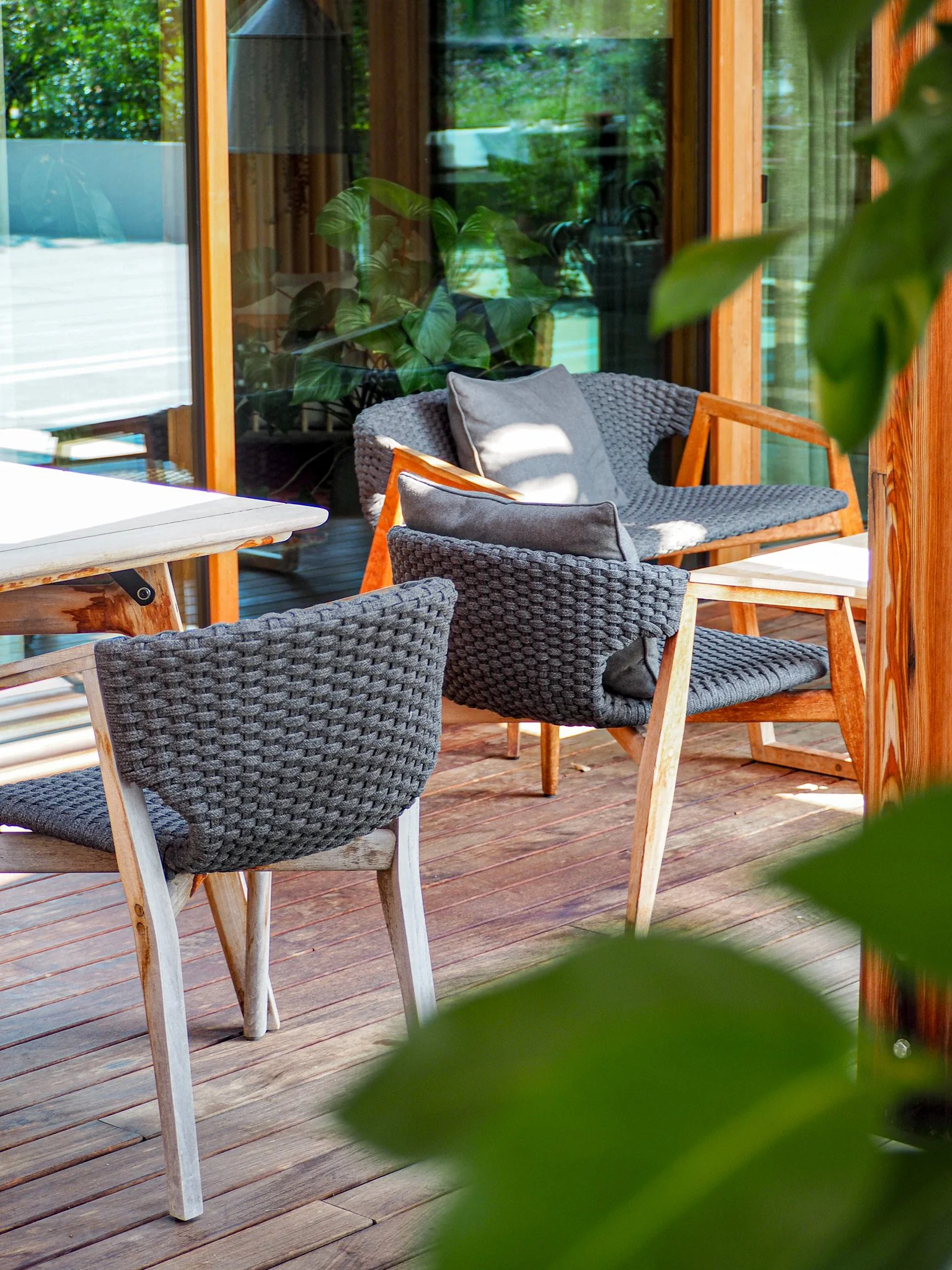 Eine Terrasse mit Holzboden, auf der mehrere Gartenstühle mit grauen Polstern und einer weißen Tischplatte stehen. Im Hintergrund sind große Fenstertüren und grüne Pflanzen sichtbar.