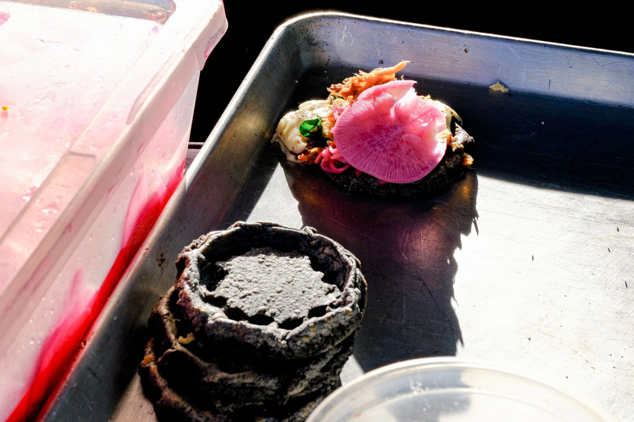 Ein Metallblech mit zwei verbrannten, schwarzen Keksen und einem bunten, großen, runden Gegenstand darauf, der aussieht wie eine Fischseite oder eine Scherbe mit rosa Muster. Im Hintergrund sind Teile einer pinken transparenten Box sichtbar.