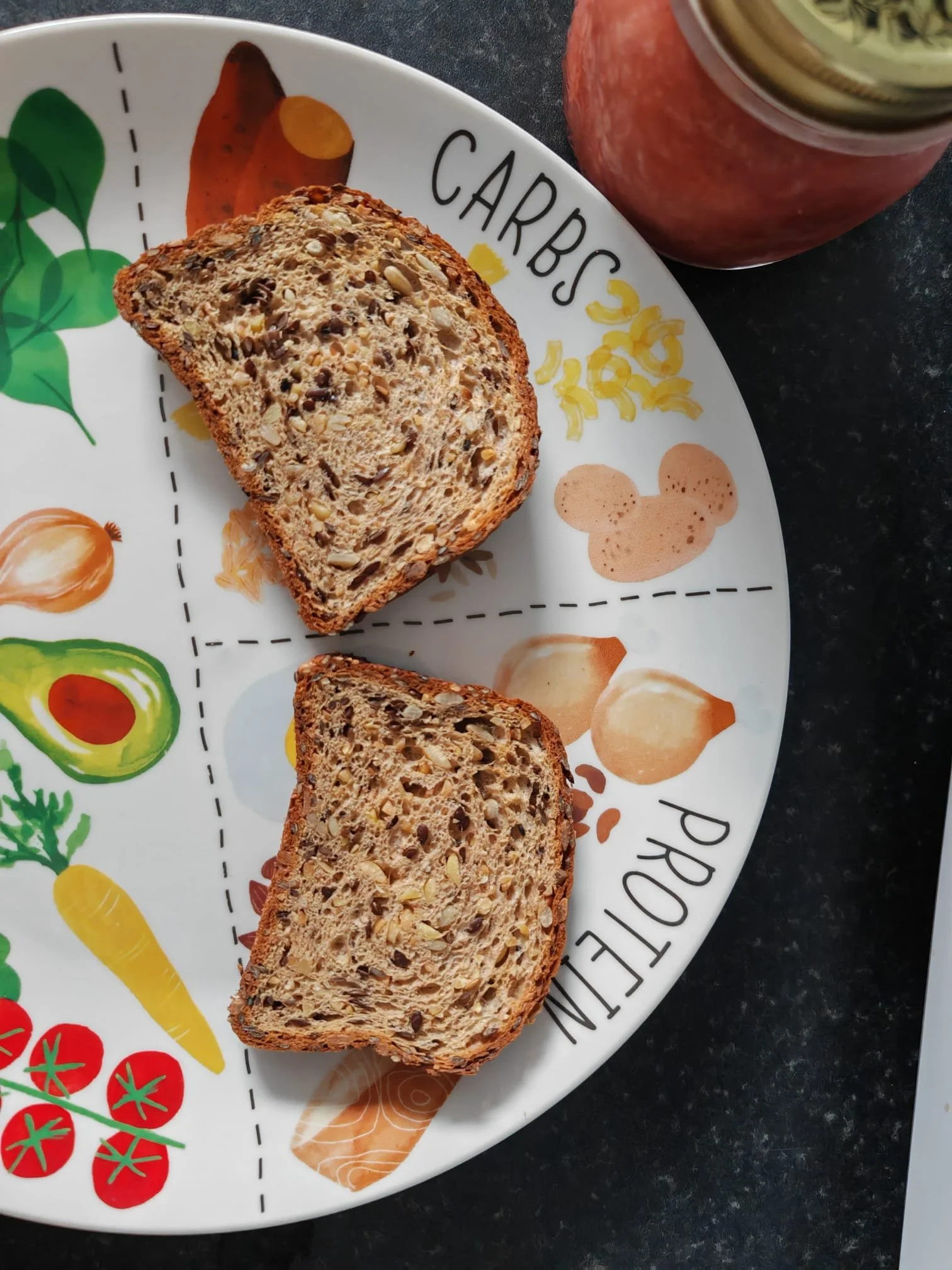 Sliced multi-seed bread on a colorful vegetable-themed plate with a jar of jam or preserves in the background.