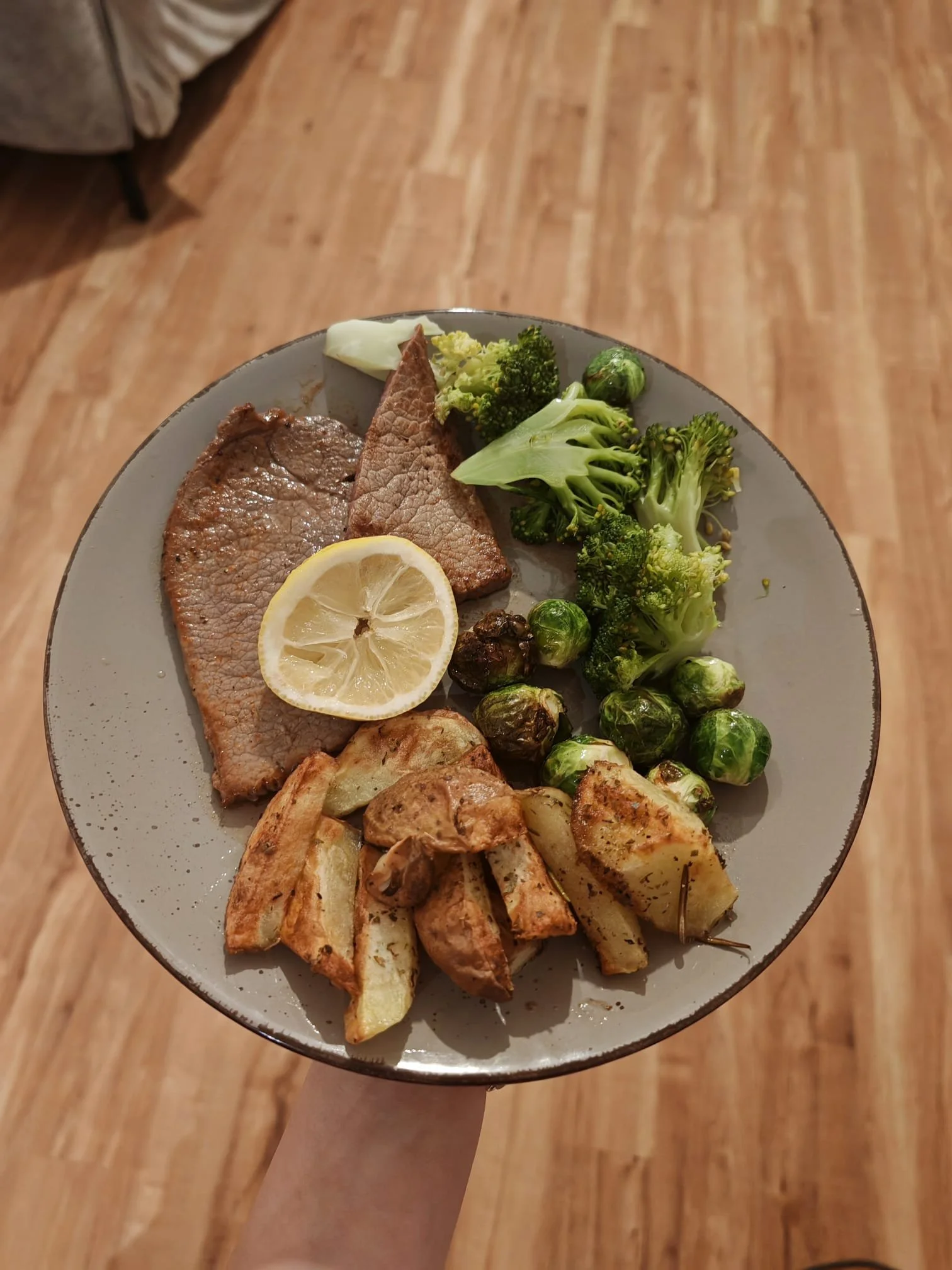 A dinner plate with sliced grilled beef, roasted potatoes, broccoli, Brussels sprouts, and a lemon wedge, held by a person's arm over a wooden floor.