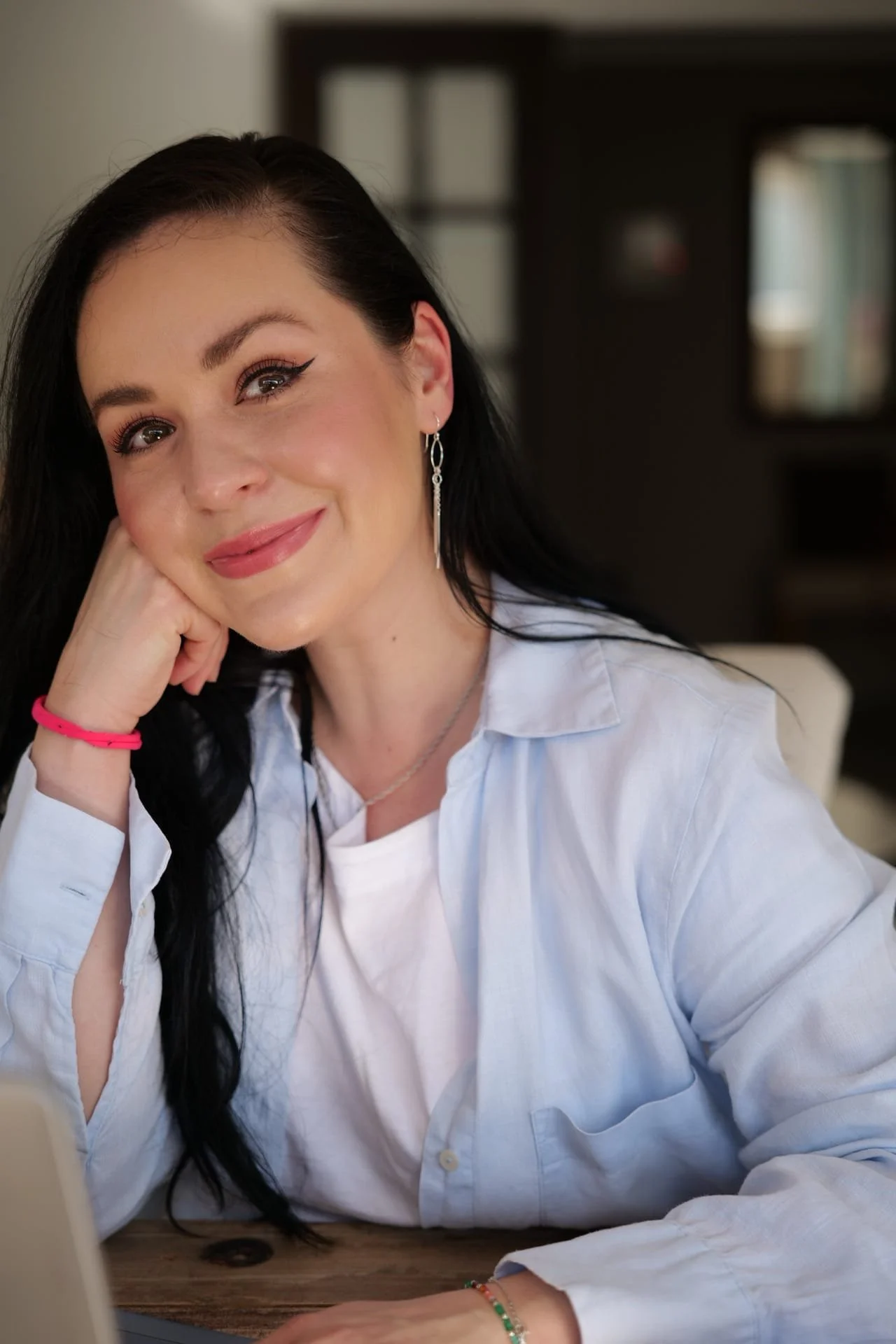 A woman with long dark hair, wearing eyeliner and earrings, is sitting at a table with her hand resting on her cheek. She is smiling and looking at the camera, wearing a white button-up shirt and a pink bracelet.