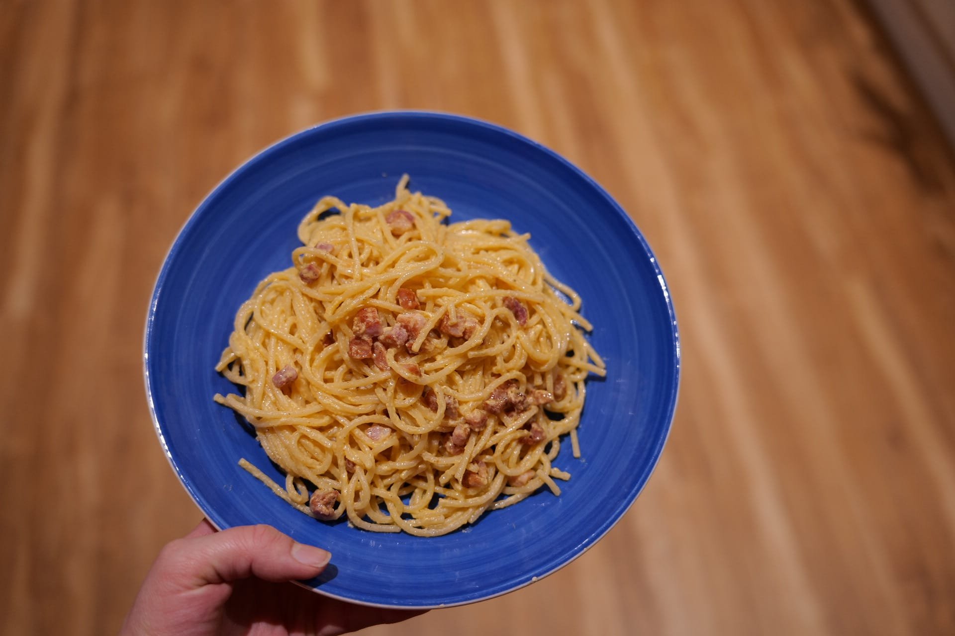 A blue bowl of spaghetti with a creamy sauce and pieces of sausage, held above a wooden surface.