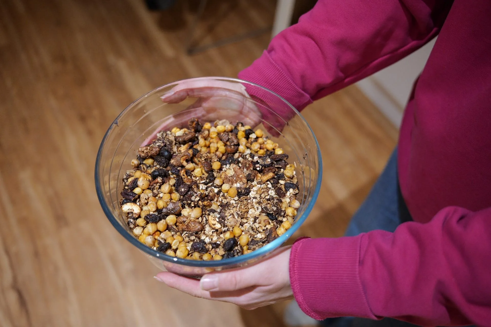 Person wearing a pink long sleeve shirt holding a glass bowl filled with mixed nuts, grains, and possibly dried fruit.