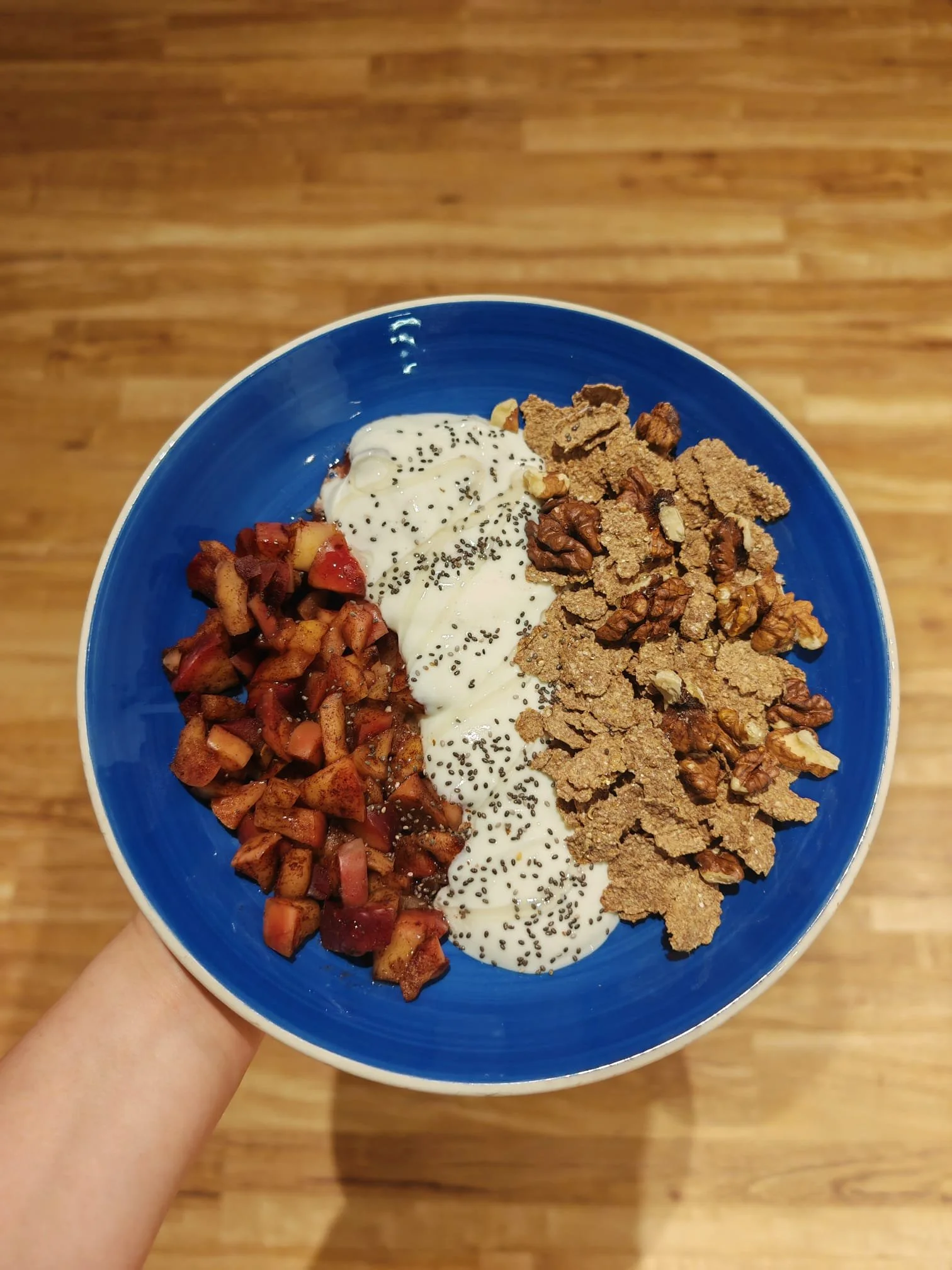 A person holding a blue bowl with diced apples, yogurt topped with chia seeds, granola, and walnuts, against a wooden floor background.
