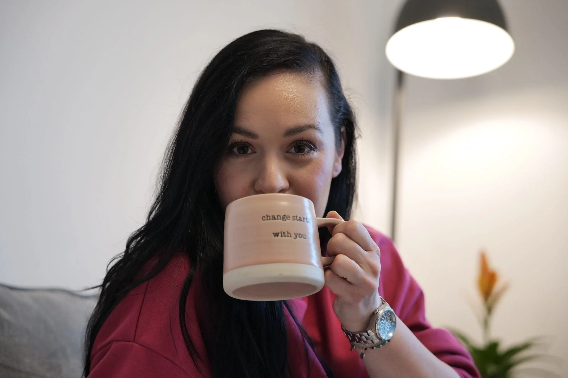 Woman with long dark hair holding a beige mug with the phrase 'change start with you' while sitting indoors near a modern floor lamp and plants.