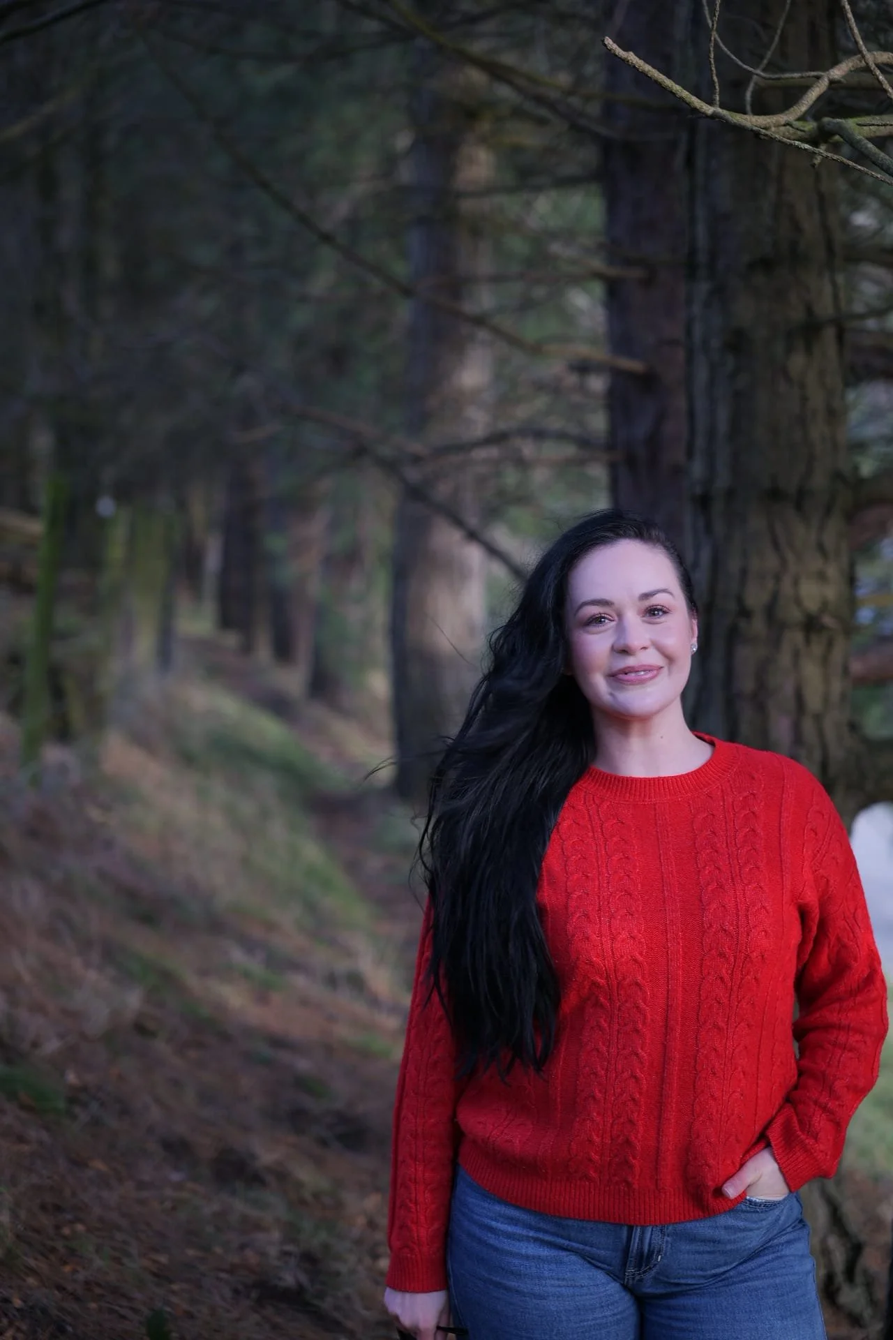 A woman with long dark hair, wearing a red knit sweater and blue jeans, standing outdoors in a wooded area with tall trees.