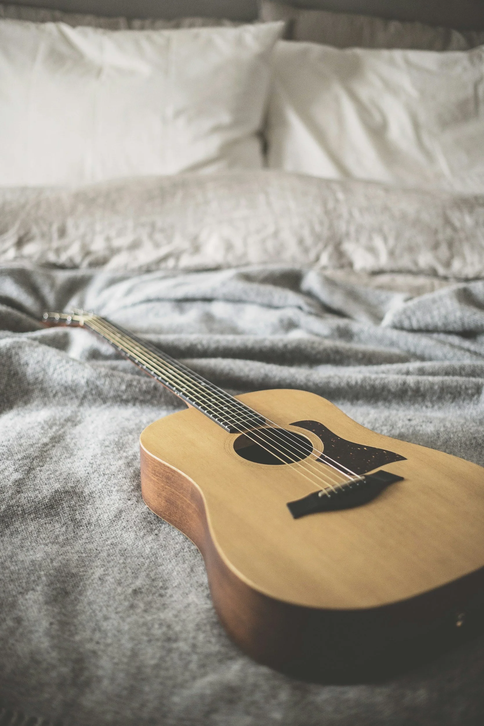An acoustic guitar resting on a gray bedspread in front of white pillows and a bed headboard.