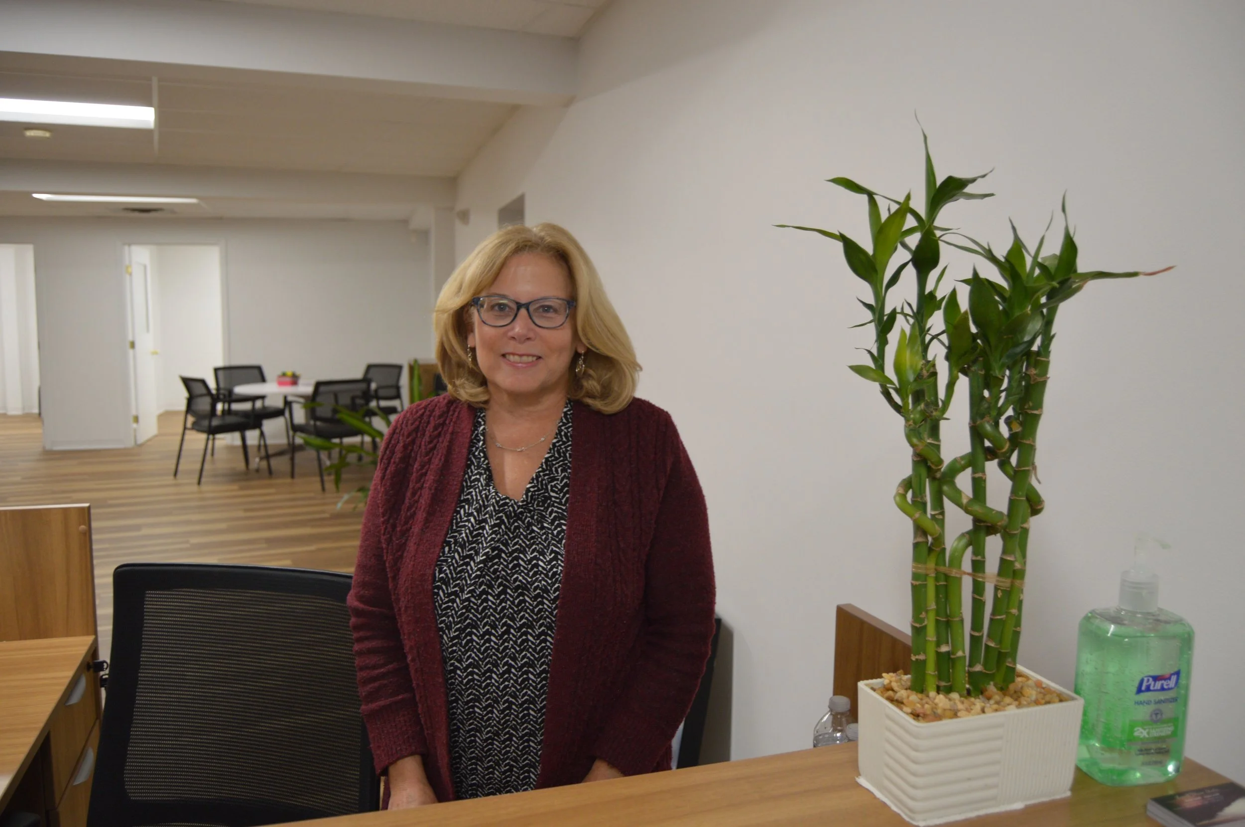 A woman with blonde hair, glasses, wearing a patterned blouse and a burgundy cardigan, standing next to a potted bamboo plant in a white rectangular planter on a wooden table in an office space.