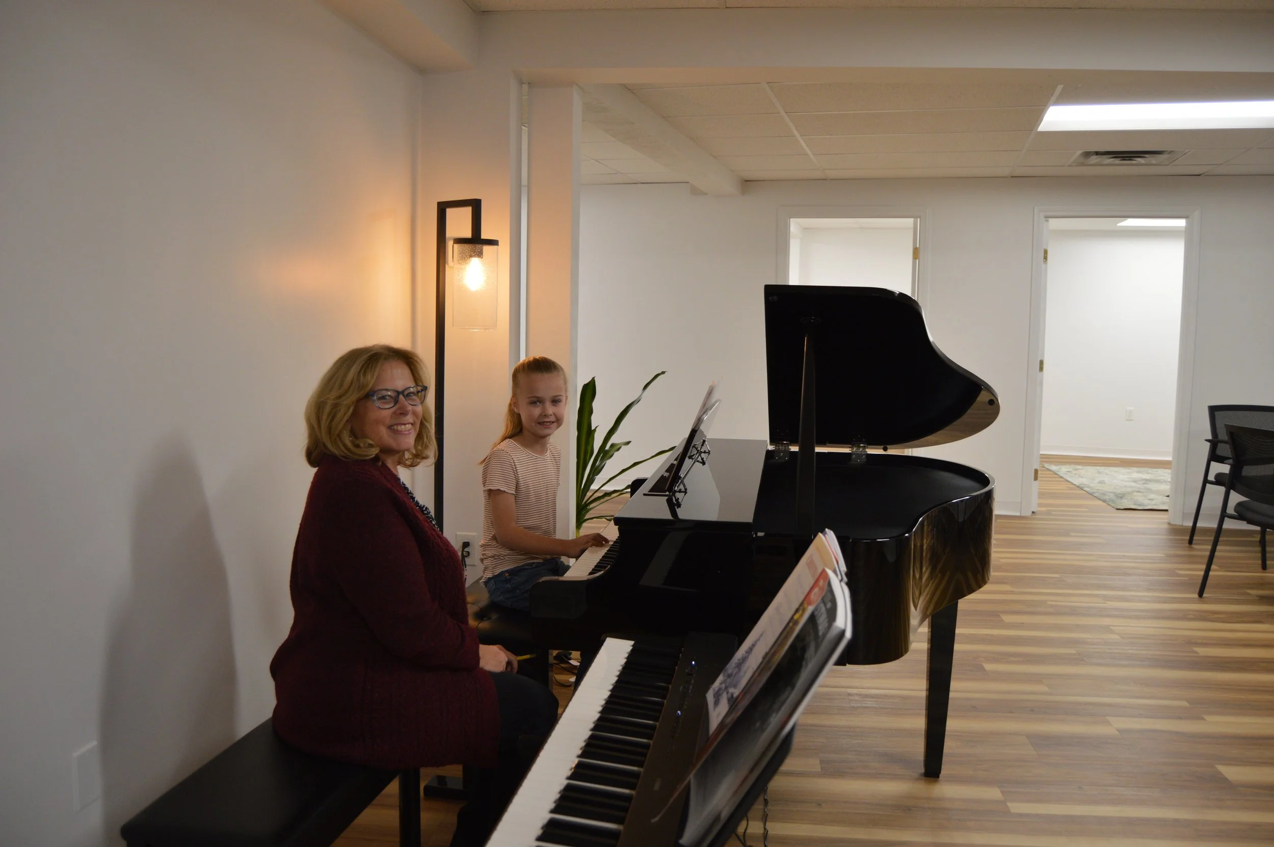 A woman and a young girl sitting at a black grand piano in a room with white walls and wooden flooring, smiling at the camera.