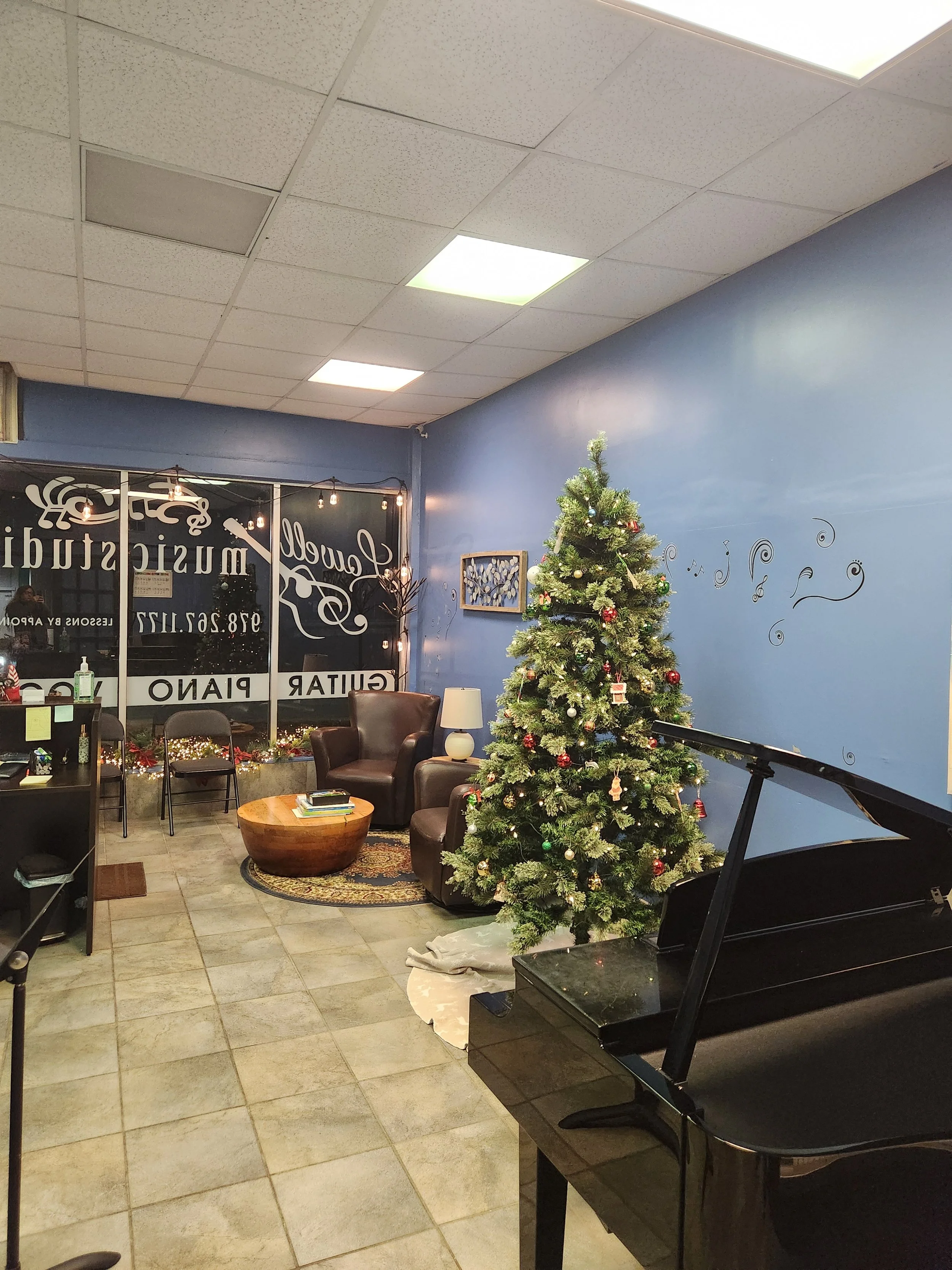 Interior of a music studio decorated for Christmas, featuring a Christmas tree with ornaments, a grand piano, a blue wall with musical notes, and a cozy seating area with chairs, a lamp, and a coffee table.