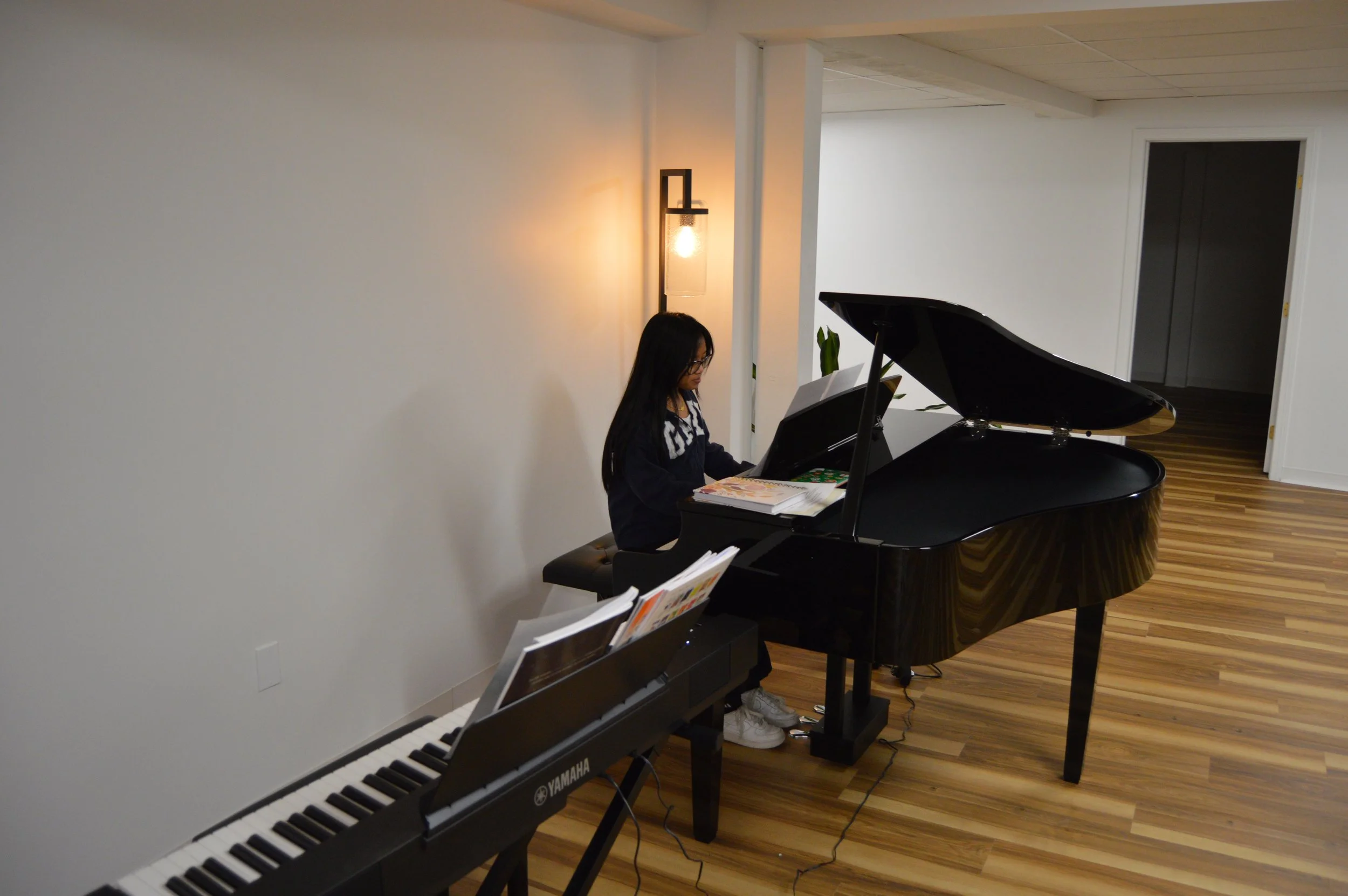 A young girl playing a black grand piano in a room with wooden floors, white walls, a wall-mounted light fixture, and a digital keyboard beside her.
