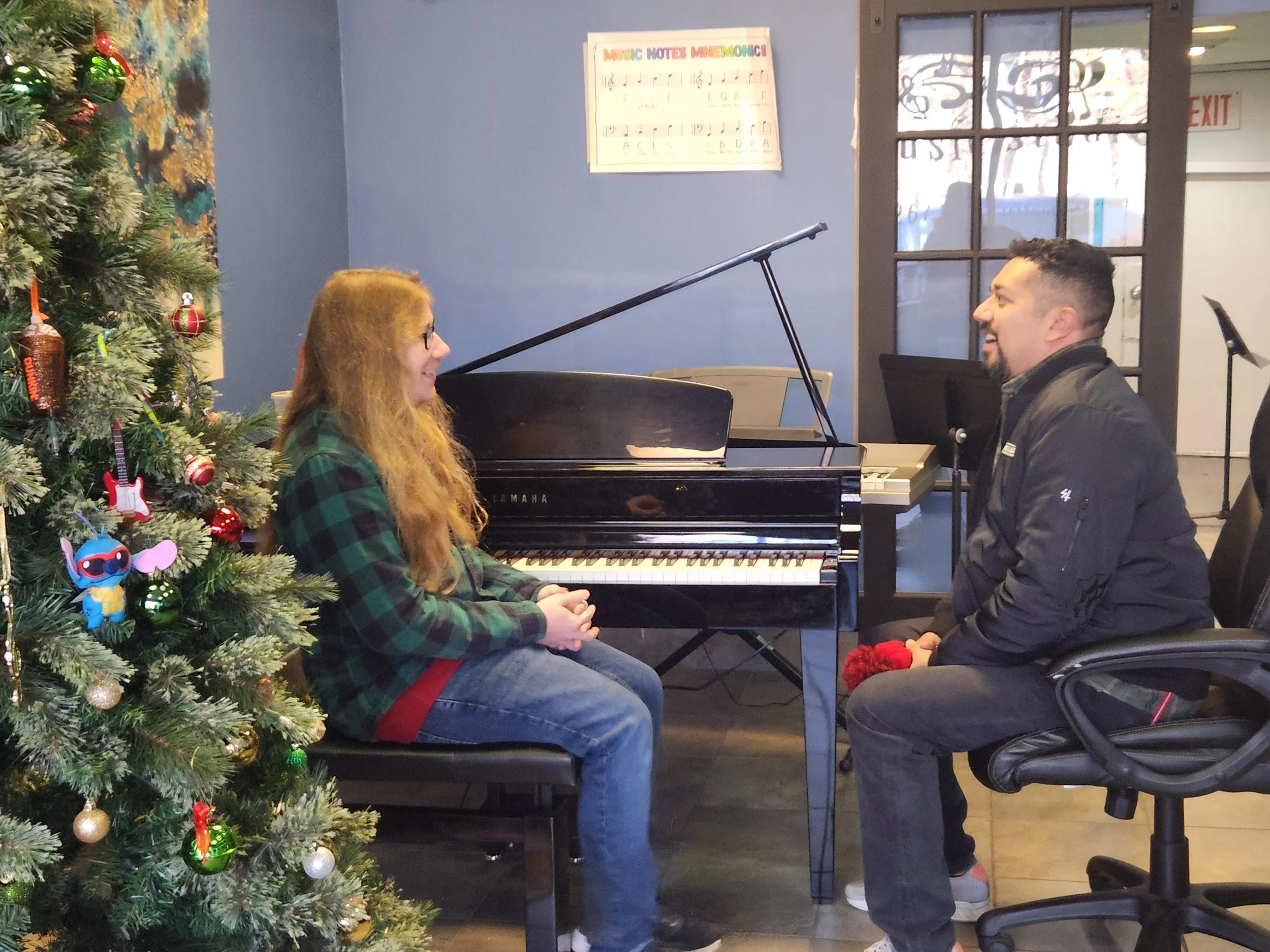 An instructor and a man sitting in front of a piano engaging in conversation in a room decorated for Christmas with a decorated Christmas tree to the left.