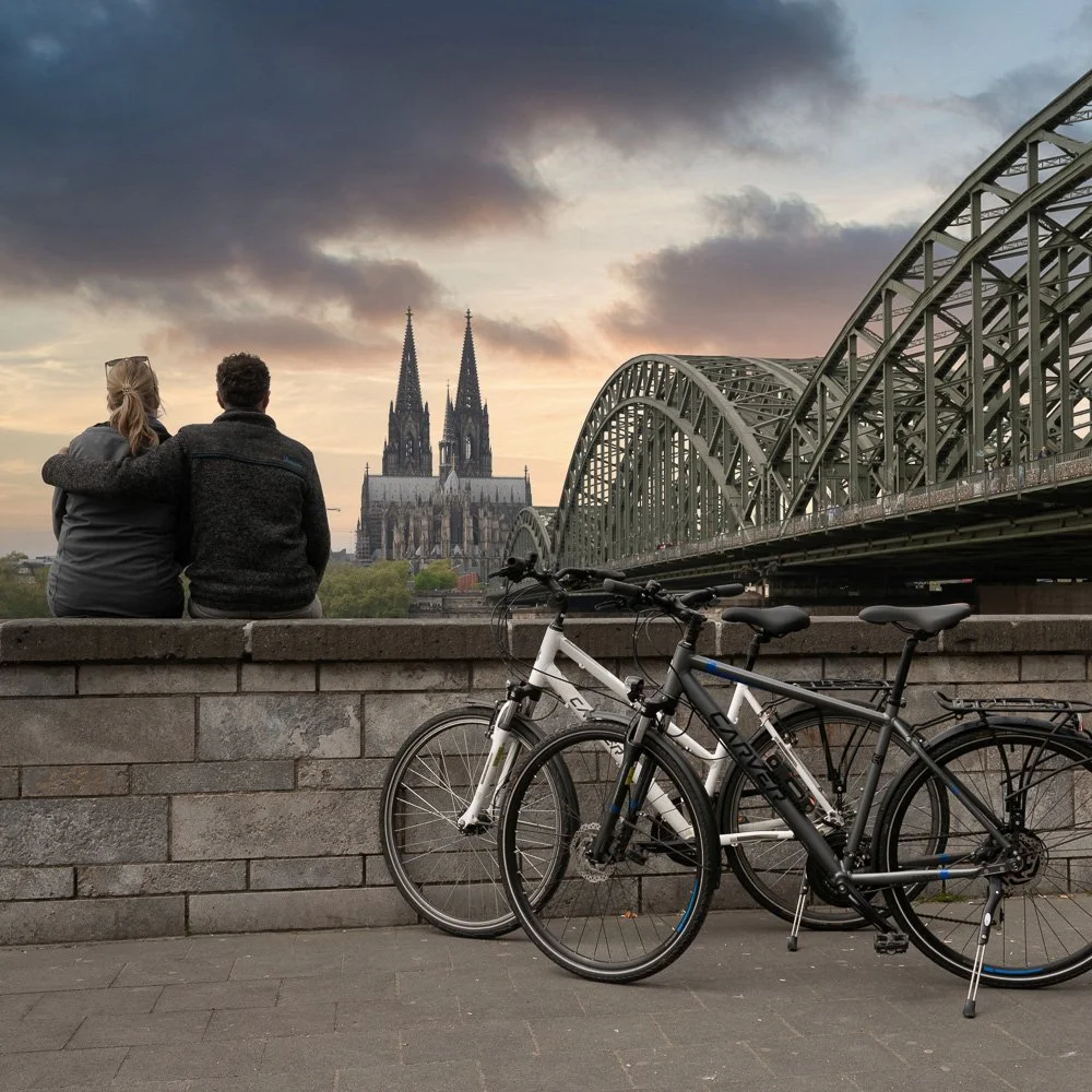 Paar an der Ufermauer vor der Hohenzollernbrücke und dem Kölner Dom im Abendlicht, Trekkingräder daneben – Tourismusfotografie in Köln