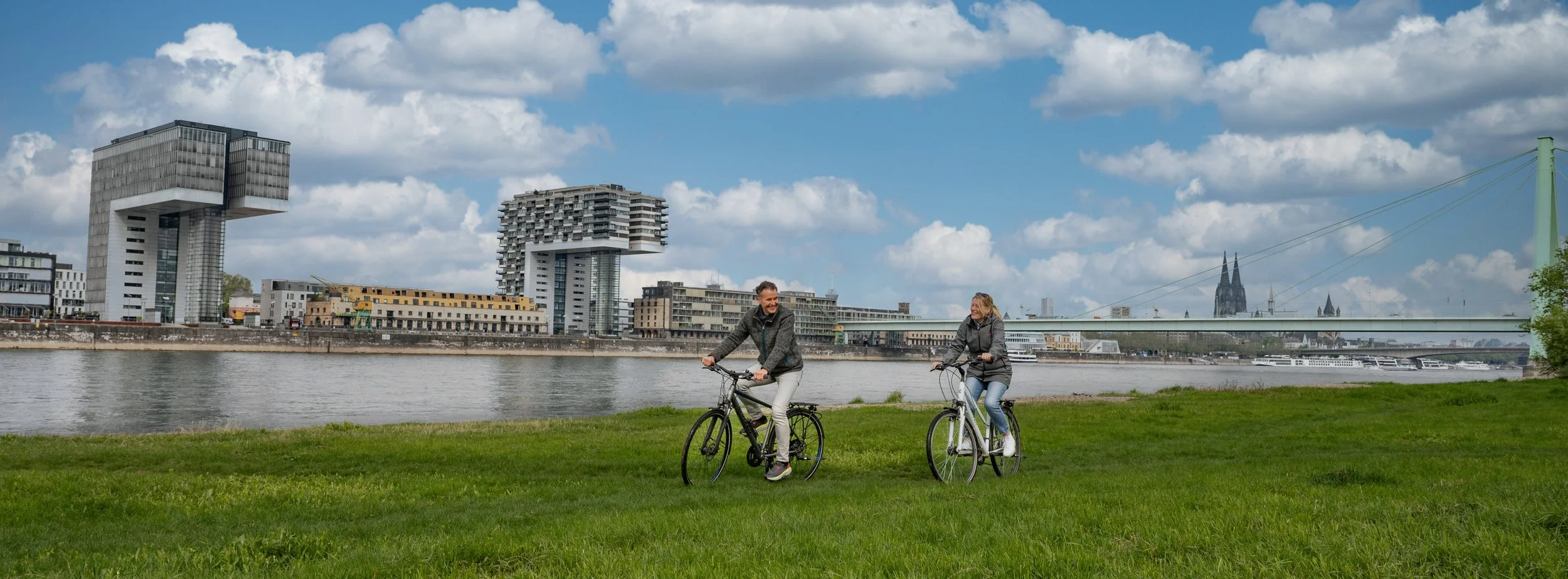Radfahrer-Paar auf der Rheinwiese vor den Kranhäusern und dem Kölner Dom – Tourismusfotografie als Panorama