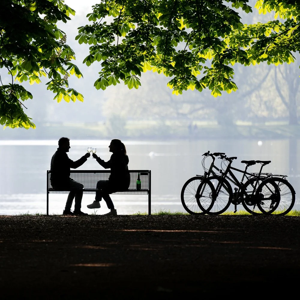 Paar stößt in der Abendstimmung auf einer Bank am Rheinufer an, Fahrräder als Silhouette – Reisefotografie in Köln