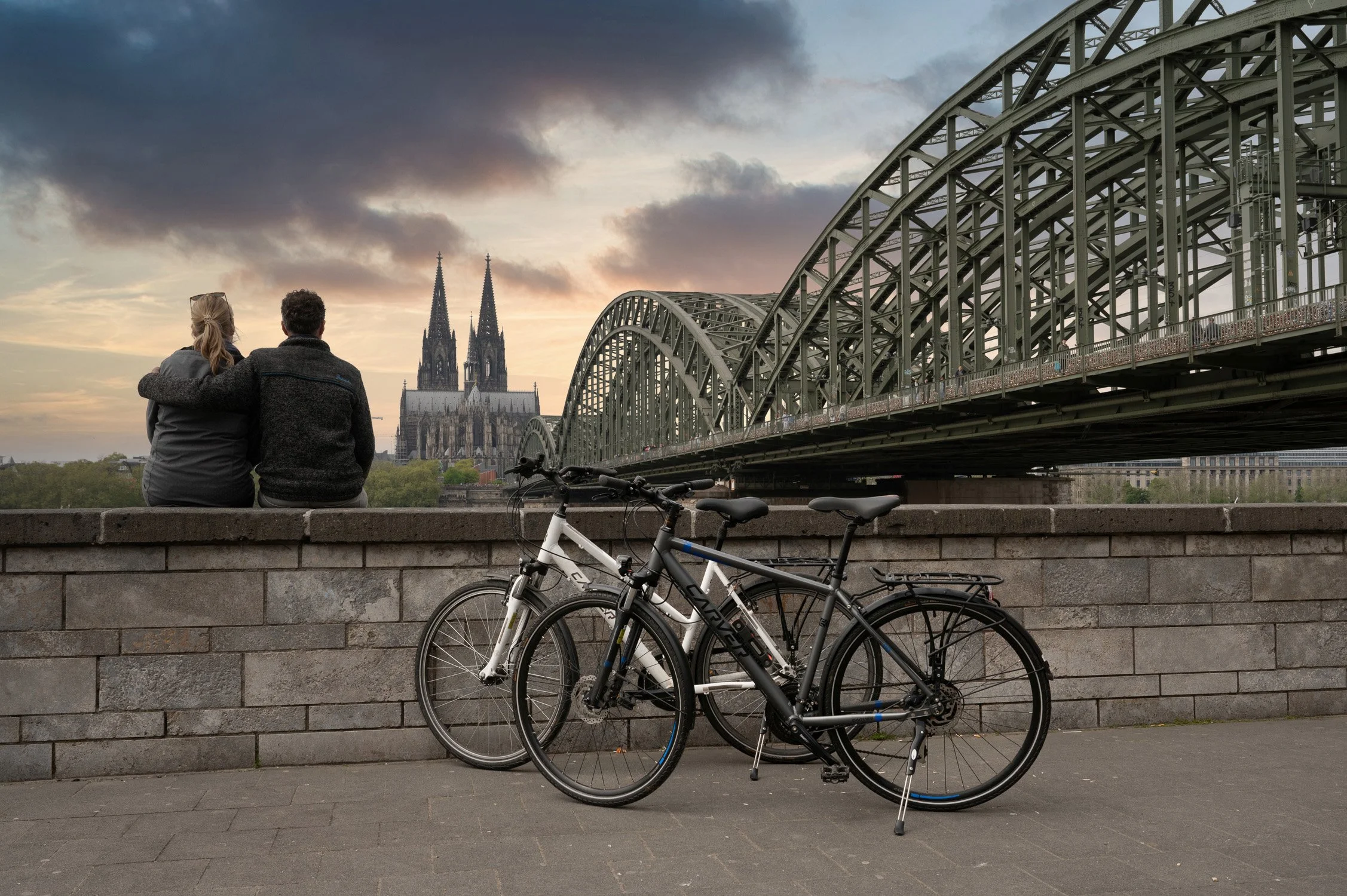 Paar an der Ufermauer vor der Hohenzollernbrücke und dem Kölner Dom im Abendlicht, Trekkingräder daneben – Tourismusfotografie in Köln